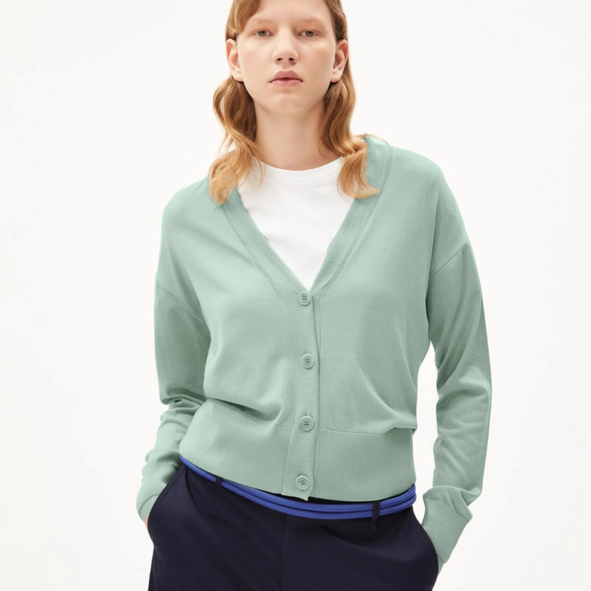 Eine Person trägt einen mintgrünen Cardigan über einem weißen T-Shirt und schwarzen Hosen vor einem hellen Hintergrund.