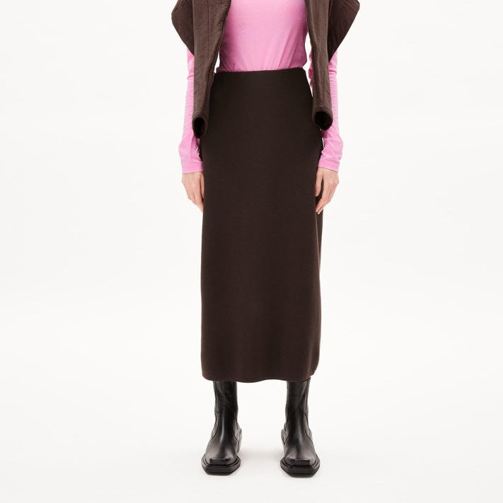 Eine Frau steht mit dem Gesicht nach vorne, trägt einen rosafarbenen Rollkragenpullover, einen braunen langen Rock, eine braune, über die Schultern gezogene Jacke, eine Strickmütze und schwarze Stiefel vor einem schlichten weißen Hintergrund.