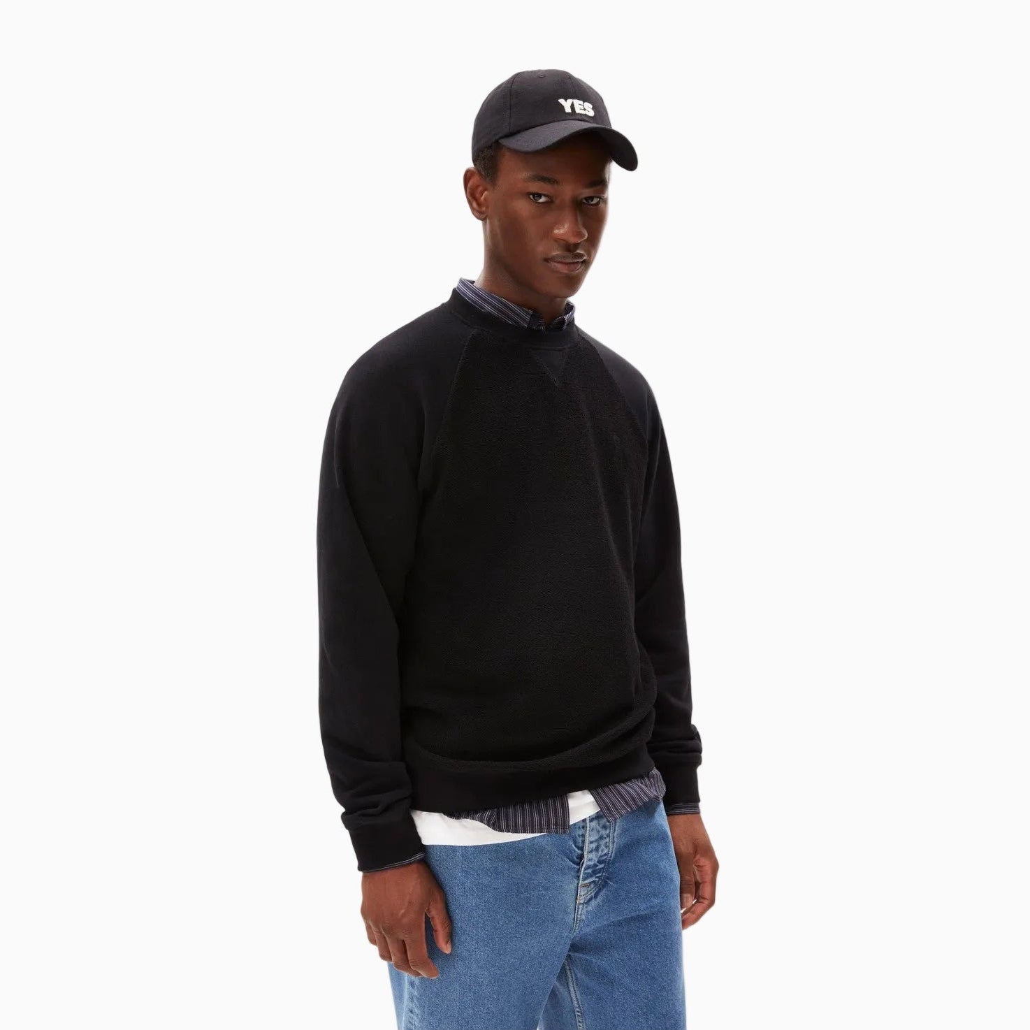Ein Mann steht mit dem Gesicht nach vorne, trägt ein schwarzes Sweatshirt über einem Hemd mit Kragen, blaue Jeans und eine schwarze Baseballmütze mit der Aufschrift YES. Der Hintergrund ist einfarbig weiß.