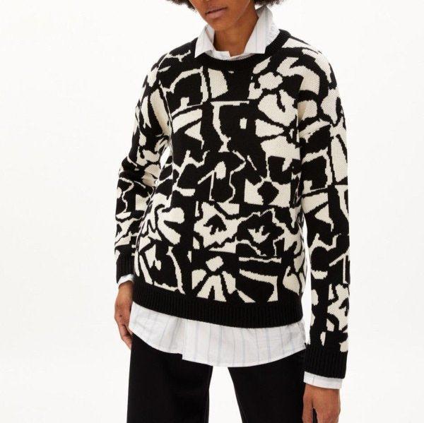 Person trägt einen schwarz-weißen Pullover mit abstraktem Muster, über einem weißen Hemd mit schwarzen Hosen.