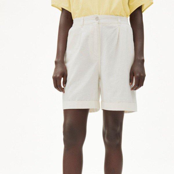 Person trägt ein hellgelbes Oberteil und weiße Shorts vor einem neutralen Hintergrund.