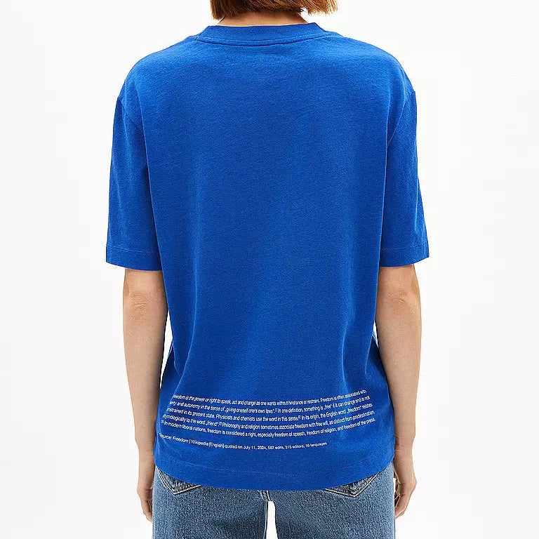 Von hinten sieht man eine Person mit kurzen Haaren, die eine cremefarbene Mütze, ein hellblaues T-Shirt mit weißer Schrift am unteren Rand und hellblaue Jeans trägt und vor einem schlichten weißen Hintergrund steht.