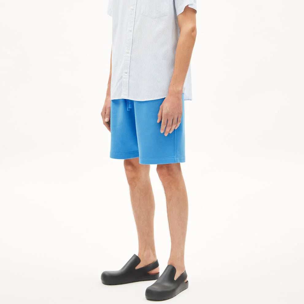 Ein Mann steht in einem weißen Raum und trägt ein hellblaues, kurzärmeliges Button-up-Hemd, hellblaue Shorts und schwarze Slipper. Er hat kurzes braunes Haar und einen neutralen Blick, der leicht zur Seite gerichtet ist.