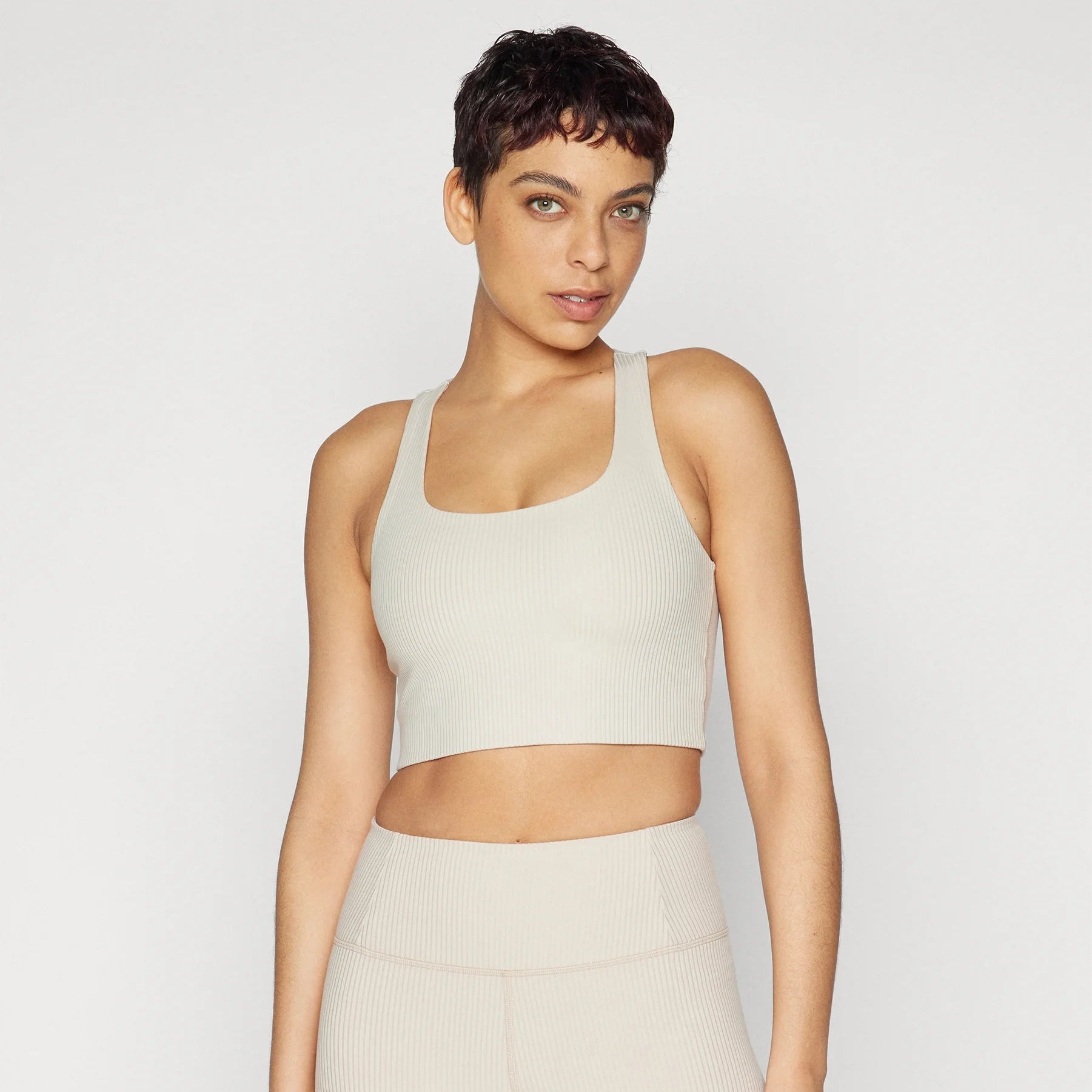 Eine Frau mit kurzen dunklen Haaren, die ein hellbeiges ärmelloses Crop-Top und dazu passende hoch taillierte Leggings trägt, steht vor einem schlichten hellen Hintergrund.