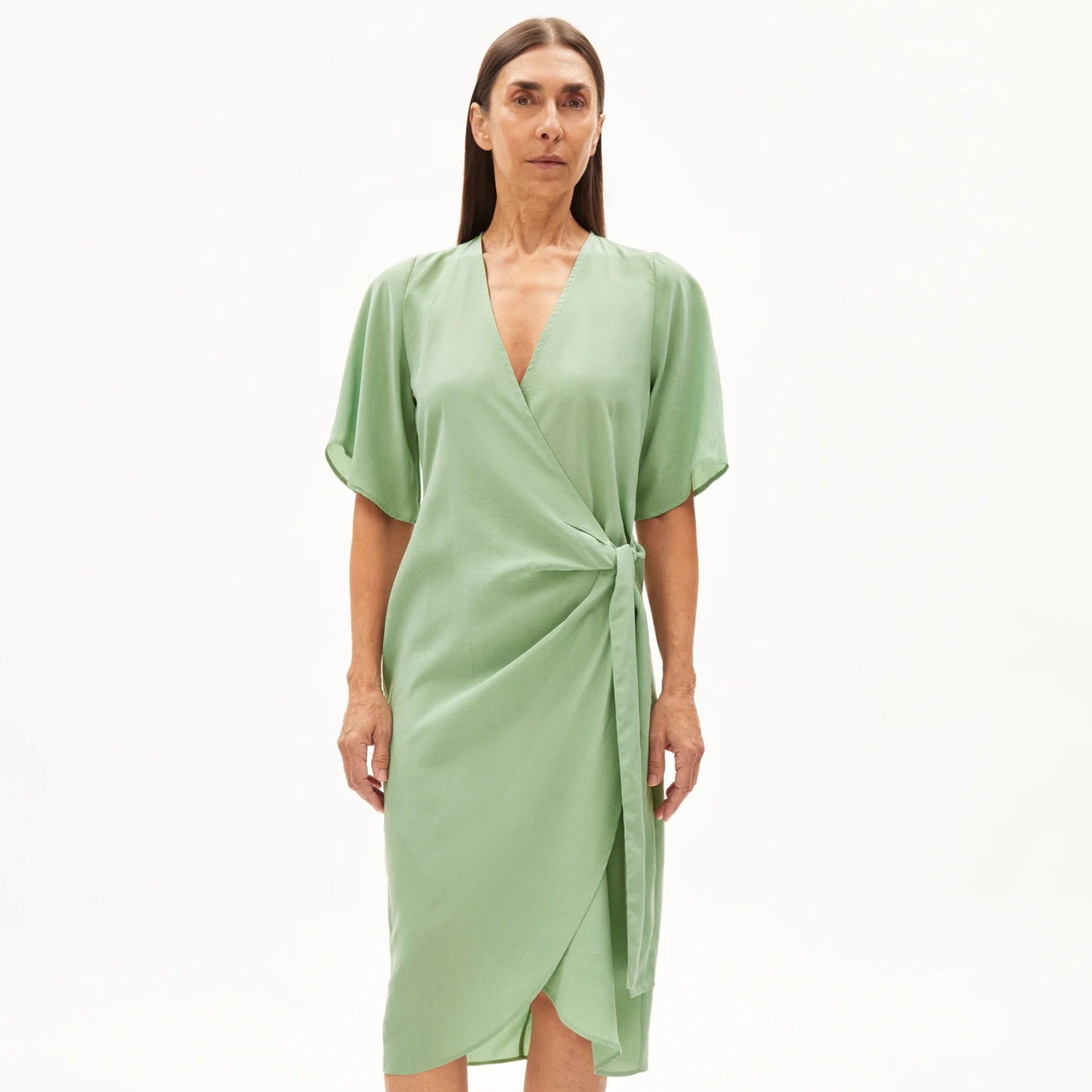 Eine Frau steht vor einem schlichten weißen Hintergrund und trägt ein hellgrünes, knielanges, kurzärmeliges Wickelkleid mit einer seitlichen Krawatte und schwarzen Riemchensandalen.