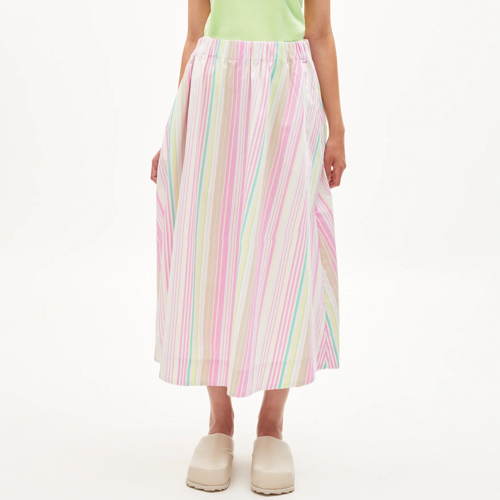Eine Frau steht vor einem weißen Hintergrund und trägt ein hellgrünes, kurzärmeliges Oberteil, einen hochtaillierten, pastellfarben gestreiften Midirock und beigefarbene Slipper.
