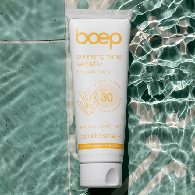 Tube Boep Sonnencreme mit LSF 30 liegt auf grünem gefliesten Boden neben Wasser mit Sonnenreflexen.