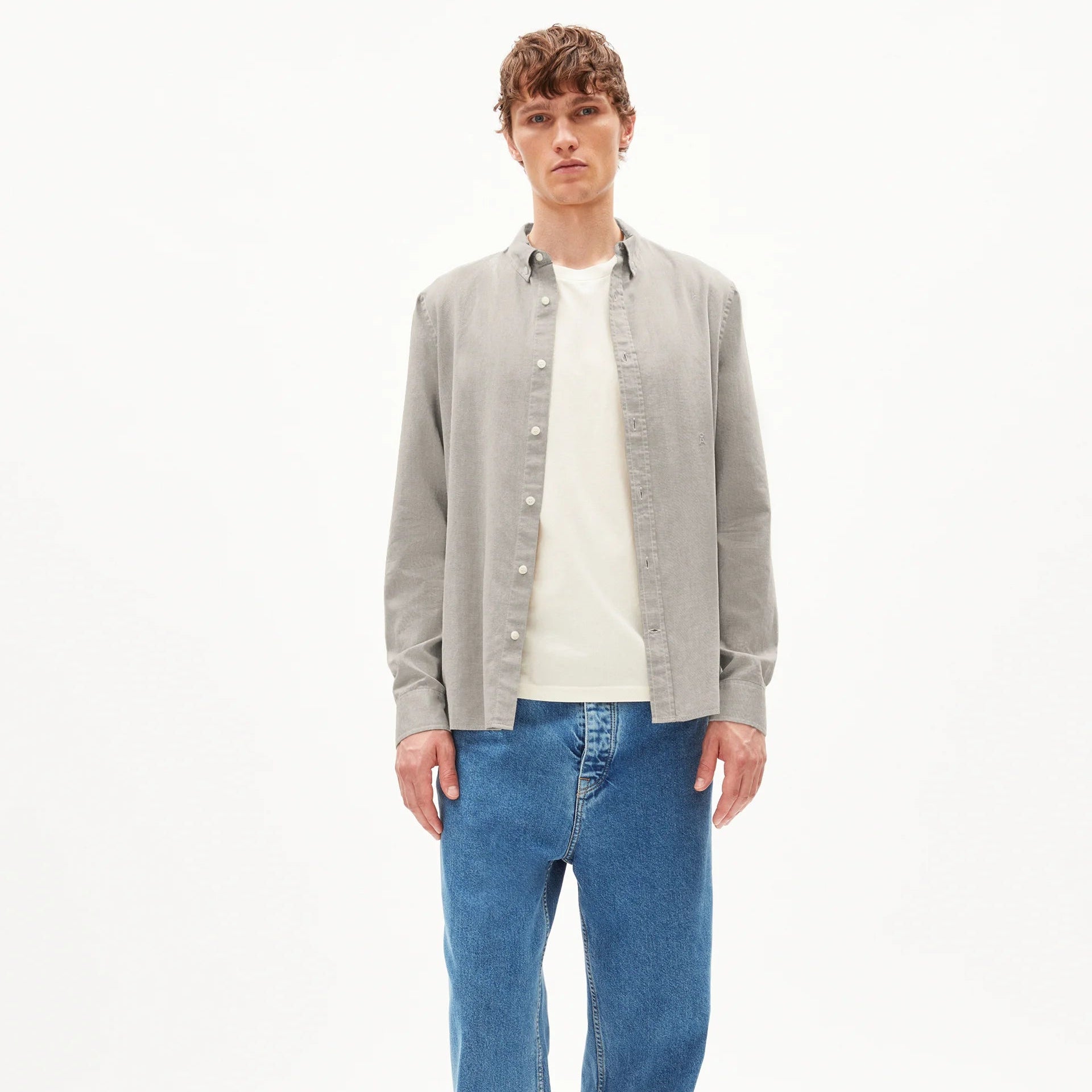 Ein junger Mann steht mit dem Gesicht nach vorne, trägt ein hellgraues Button-up-Hemd über einem weißen T-Shirt, blaue Jeans und beigefarbene Schuhe vor einem schlichten weißen Hintergrund.