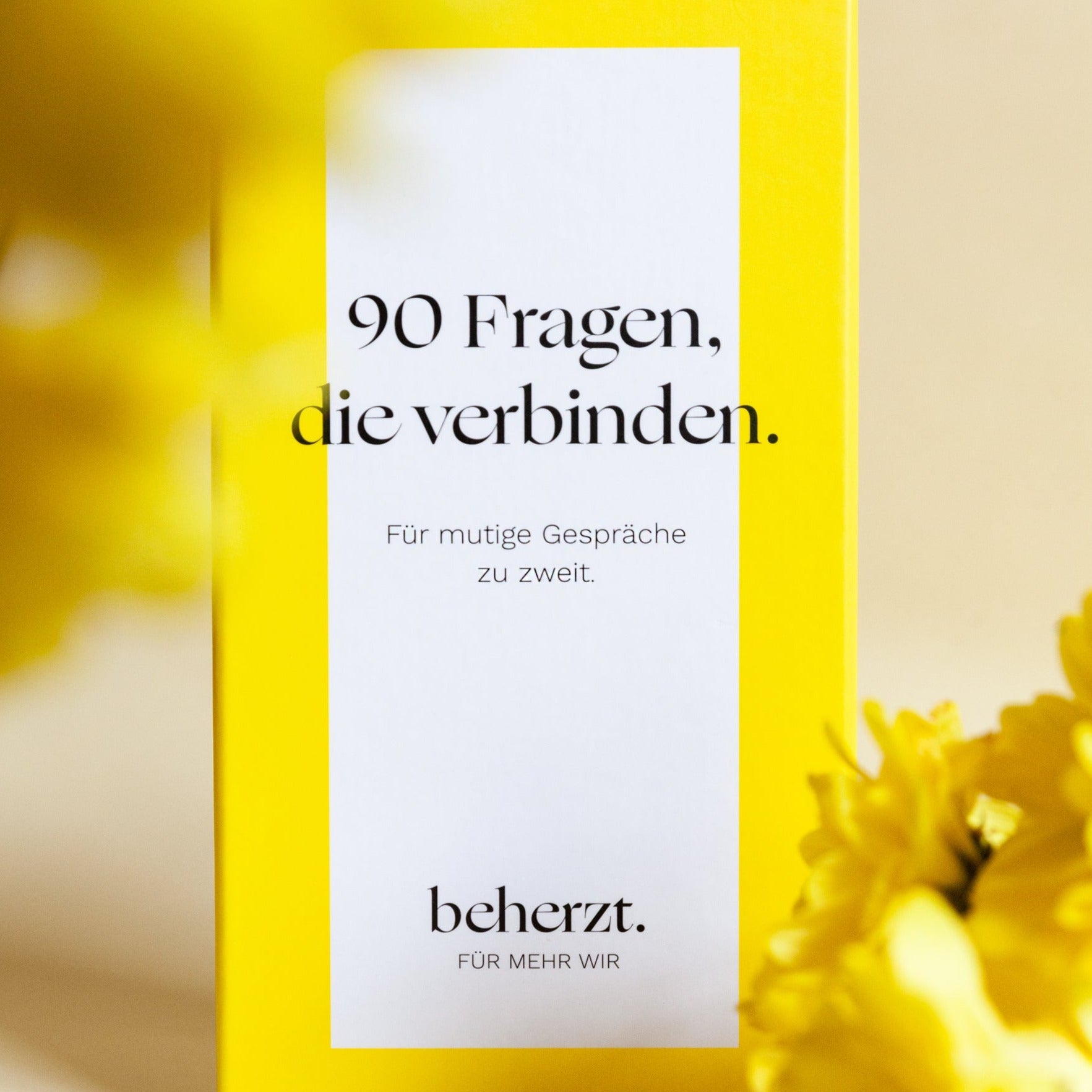Gelbe Verpackung mit Text "90 Fragen, die verbinden. Für mutige Gespräche zu zweit." und gelben Blumen im Vordergrund.