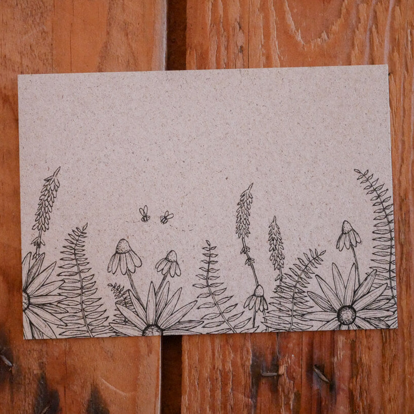 Eine handgezeichnete Illustration von Wildblumen und Farnen mit zwei Bienen auf einem Blatt hellbraunem Papier, das auf eine rustikale Holzfläche gepinnt ist.