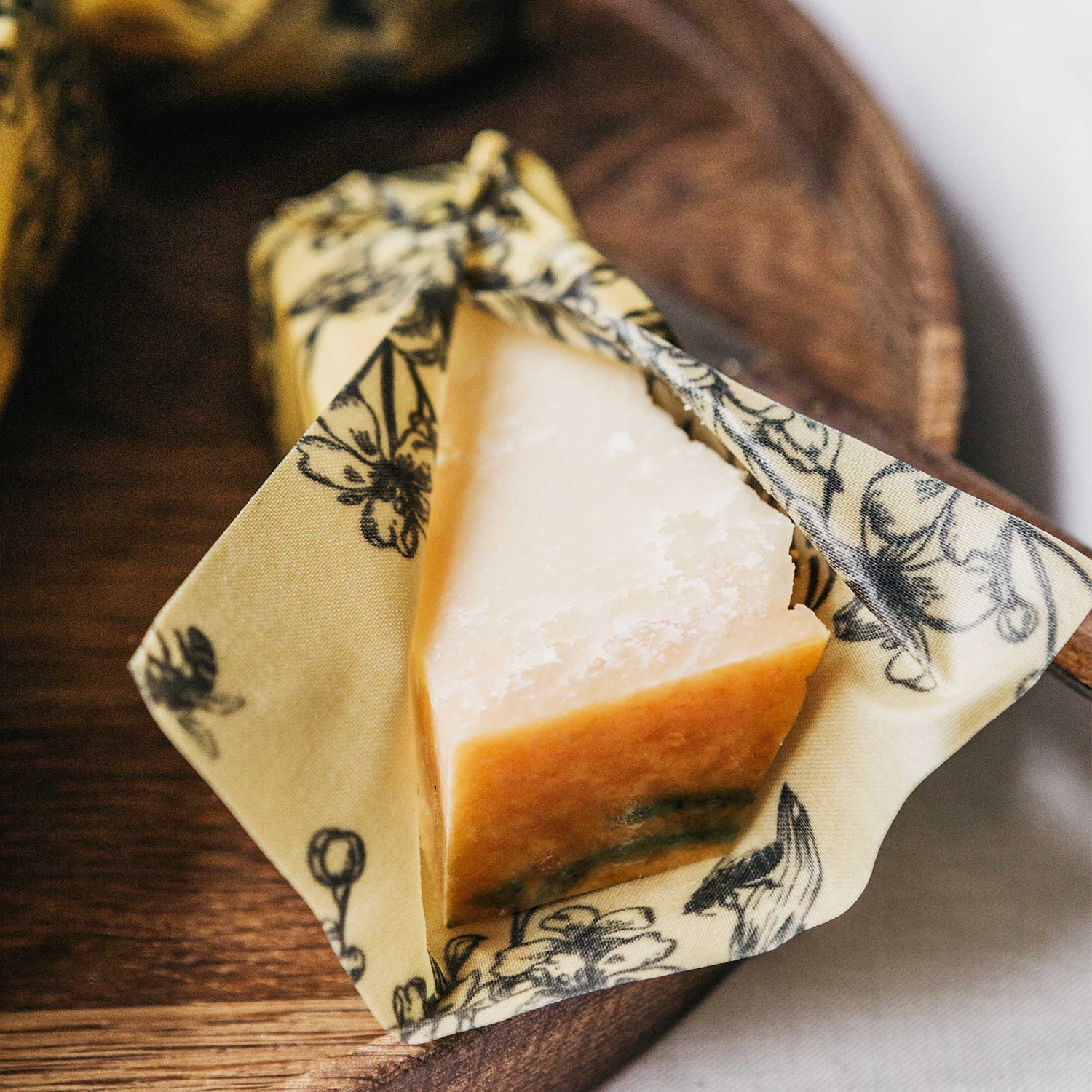 Ein Stück Hartkäse ist in einem Bienenwachstuch mit Blumendruck auf einem Holzbrett verpackt.