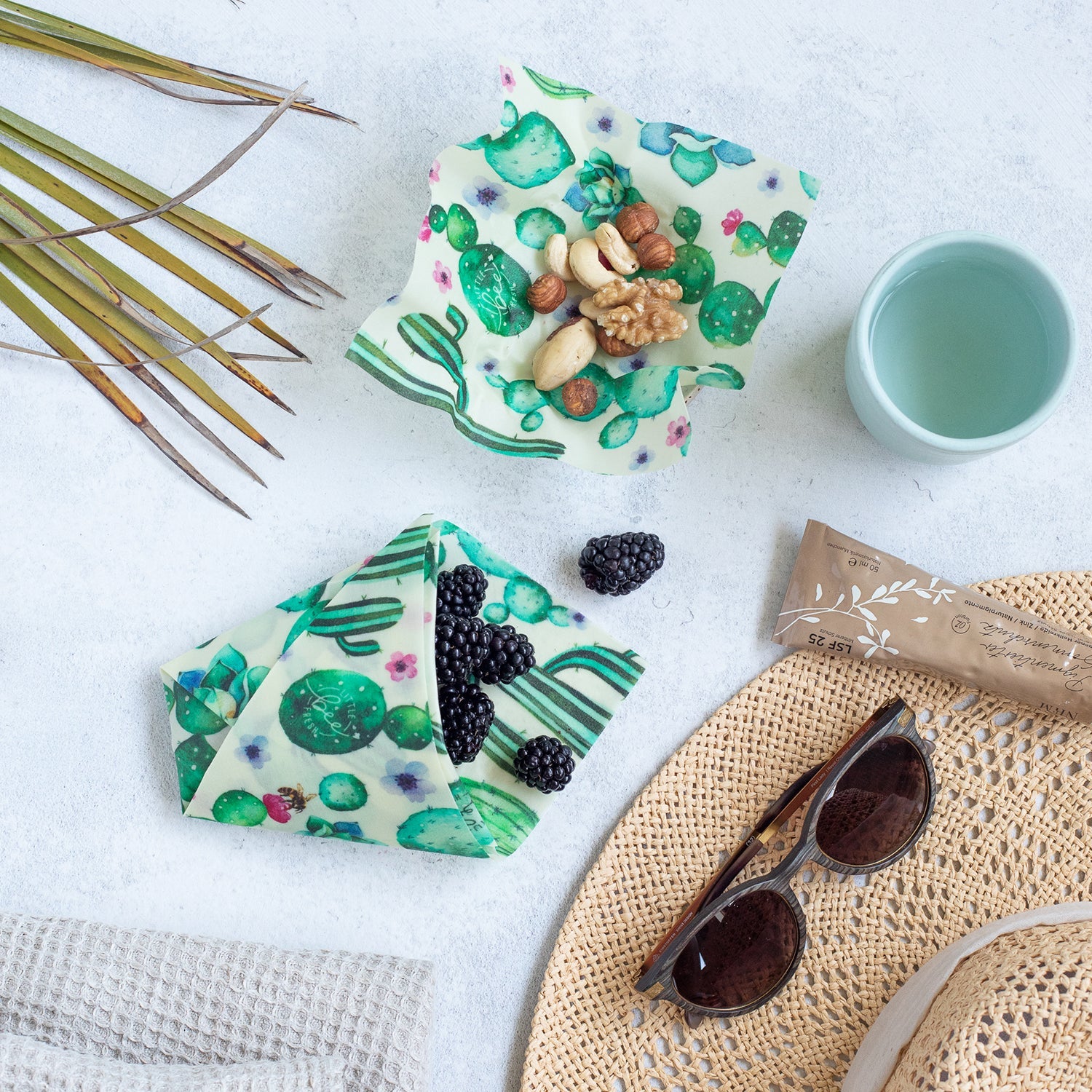 Nüsse, Beeren und ein Getränk neben einer Sonnenbrille auf einem Strohhut und Stofftücher mit Kaktusmuster.