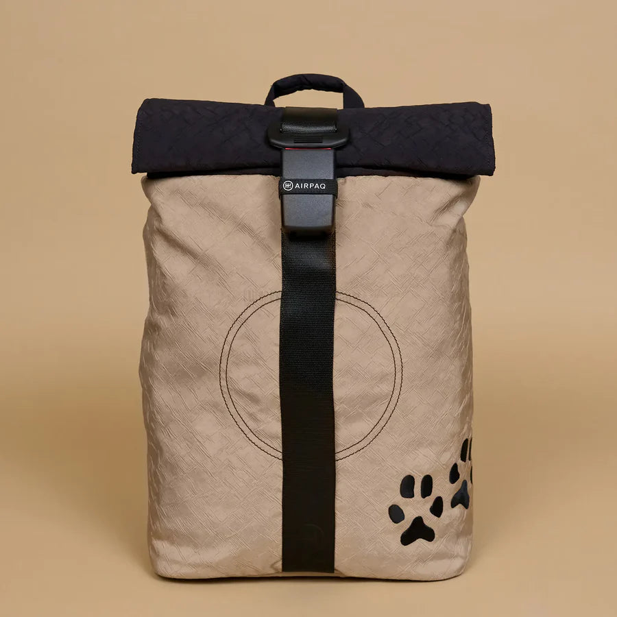 Ein beiger Rolltop-Rucksack mit schwarzem Riemen, schwarzer Klappe, kreisförmiger Naht auf der Vorderseite und einem kleinen schwarzen Pfotenabdruck in der unteren rechten Ecke vor einem einfarbigen hellbraunen Hintergrund.