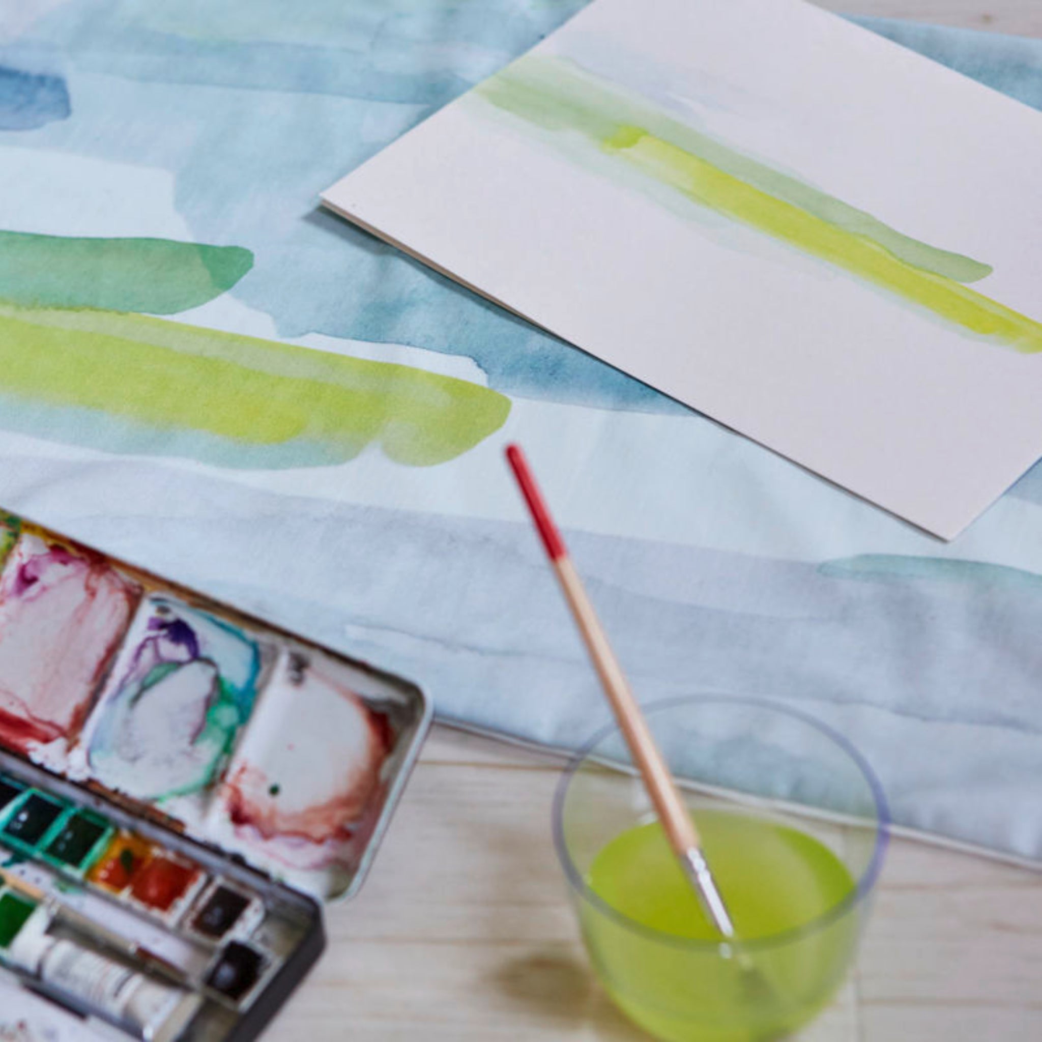Ein Aquarellfarbkasten, ein Pinsel in einem Glas Wasser und ein bemaltes Blatt Papier auf einem Tisch.