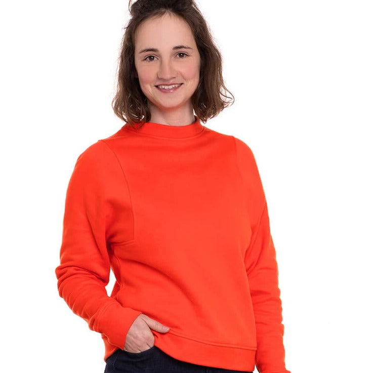 Person in vibrant orange sweater smiling with hands in pockets against a white background.