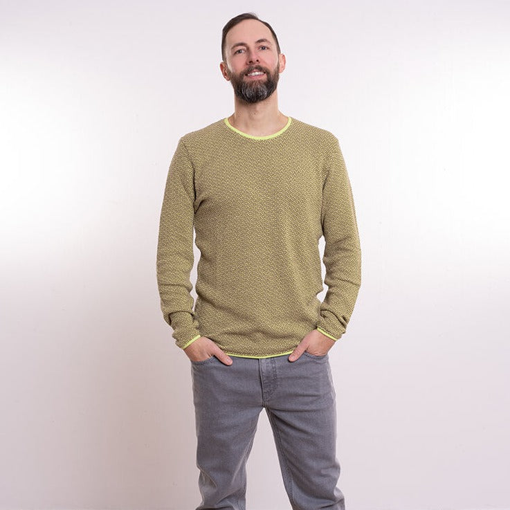 A man with a beard stands in a light olive sweater and gray pants against a plain white background, smiling.