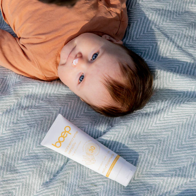 Ein Baby liegt auf einer Decke mit Sonnenschutzcreme auf der Nase, neben einer Tube Sonnencreme.