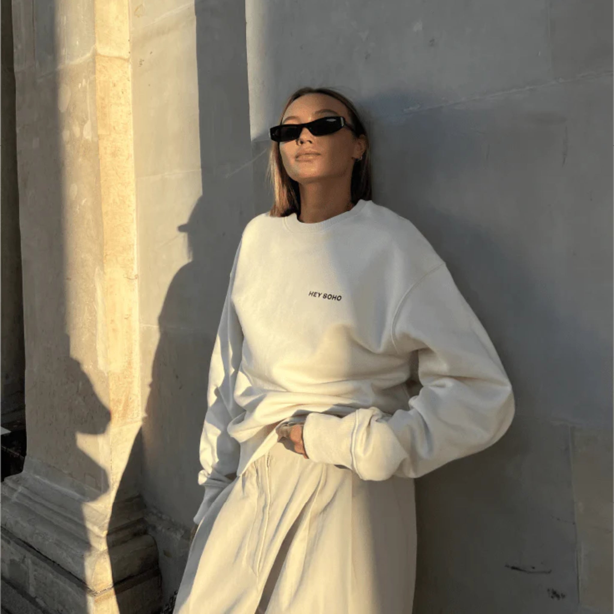 Person in a white outfit leans against a stone wall, wearing dark sunglasses and standing in sunlight.