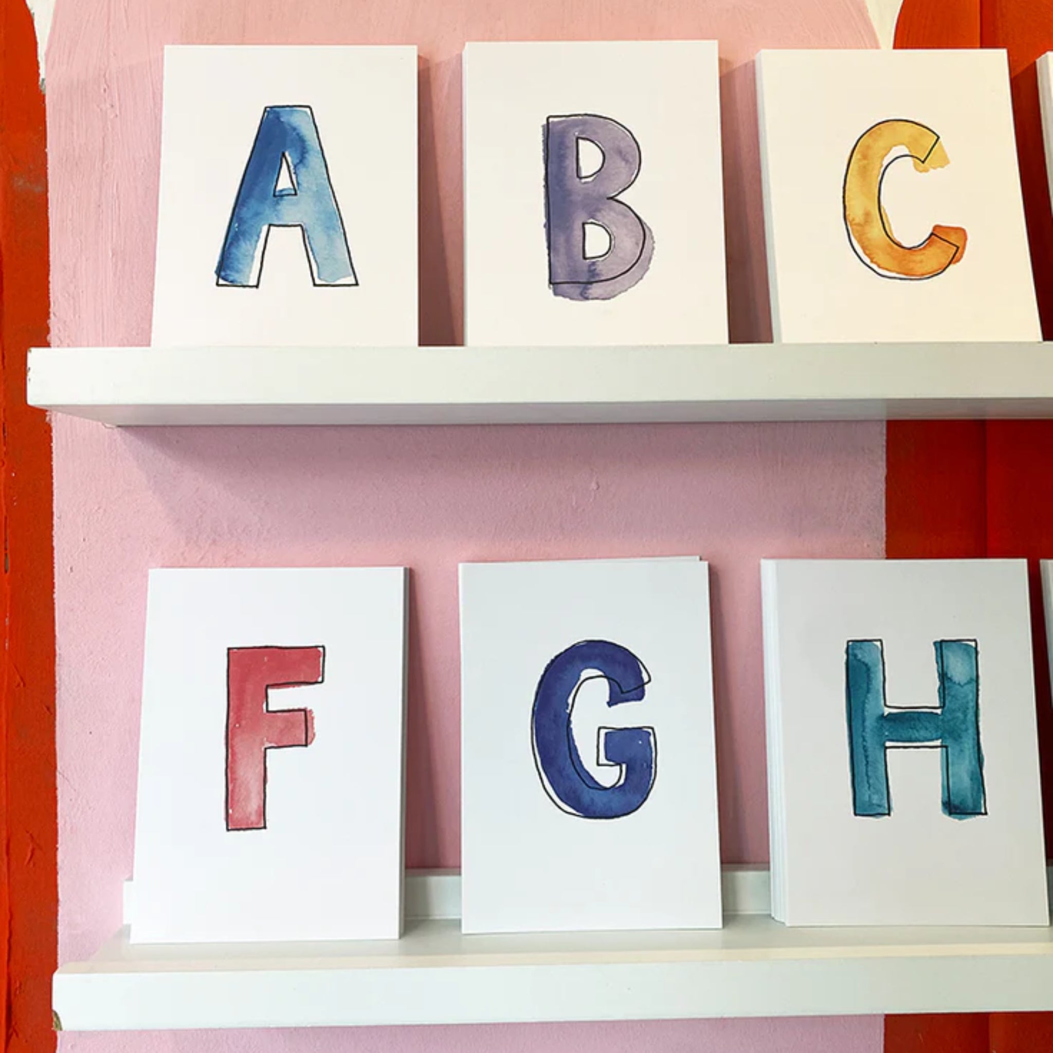 Weiße Karten mit handgemalten Buchstaben A, B, C, F, G und H in bunten Aquarellfarben auf Regalen vor rosa Wand.