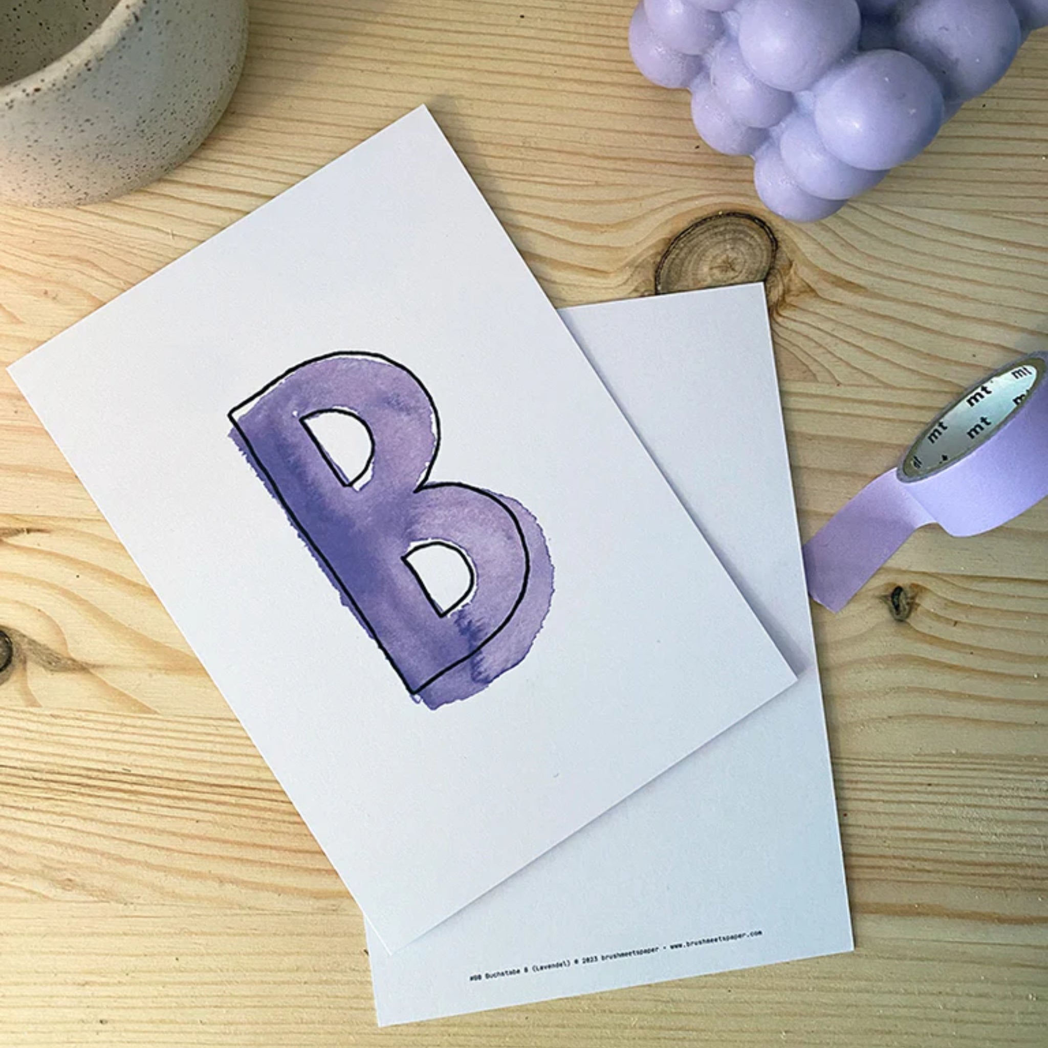 Eine lilafarbene Karte mit dem Buchstaben "B" liegt auf einem Holztisch neben lila Klebeband und einer Kerze.