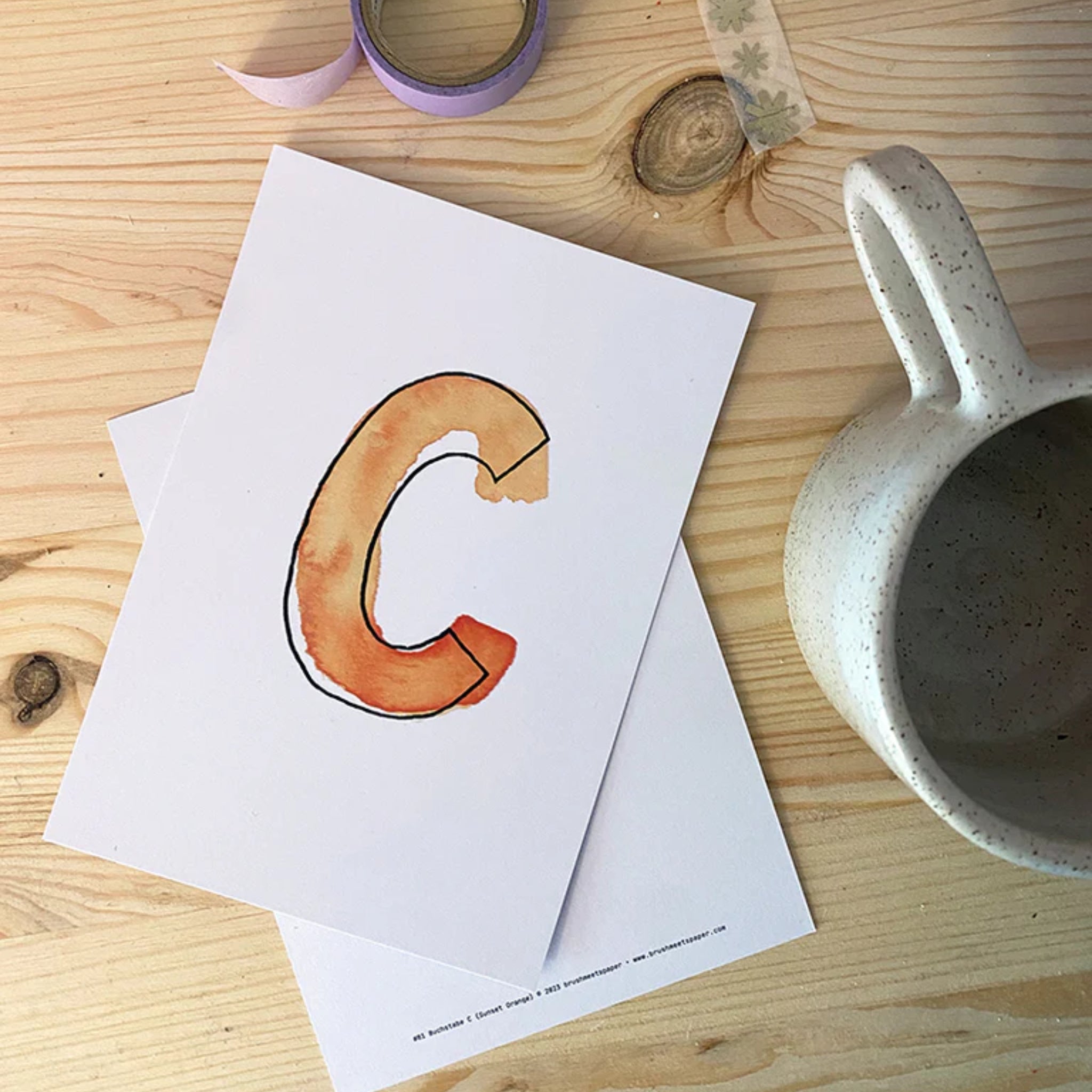 Aquarellkarte mit einem stilisierten "C" auf einem Holztisch, daneben eine graue Tasse und bunte Klebebänder.