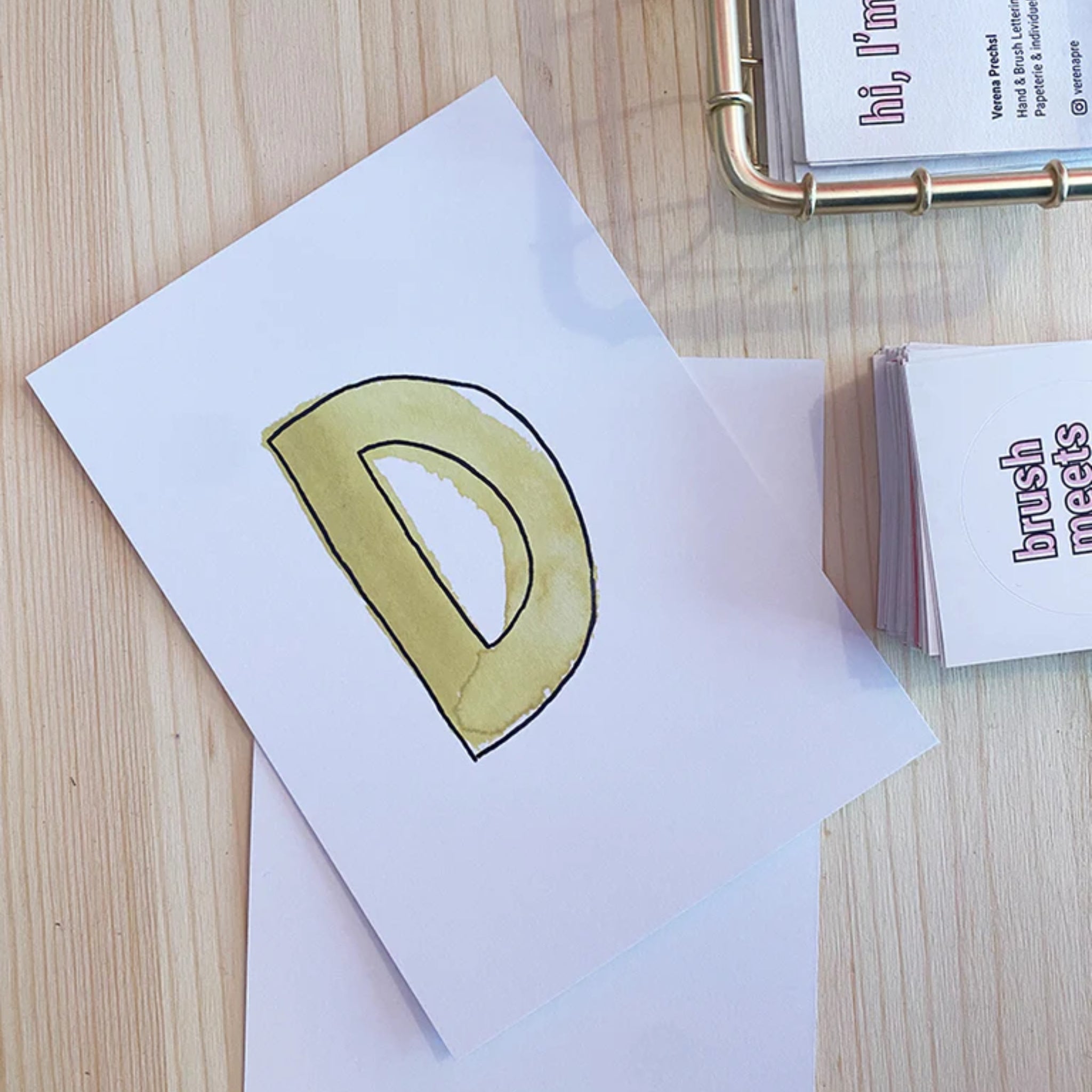 Eine Karte mit einem großen, handgemalten Buchstaben "D" in Gelb liegt auf einem Holztisch neben Visitenkarten.