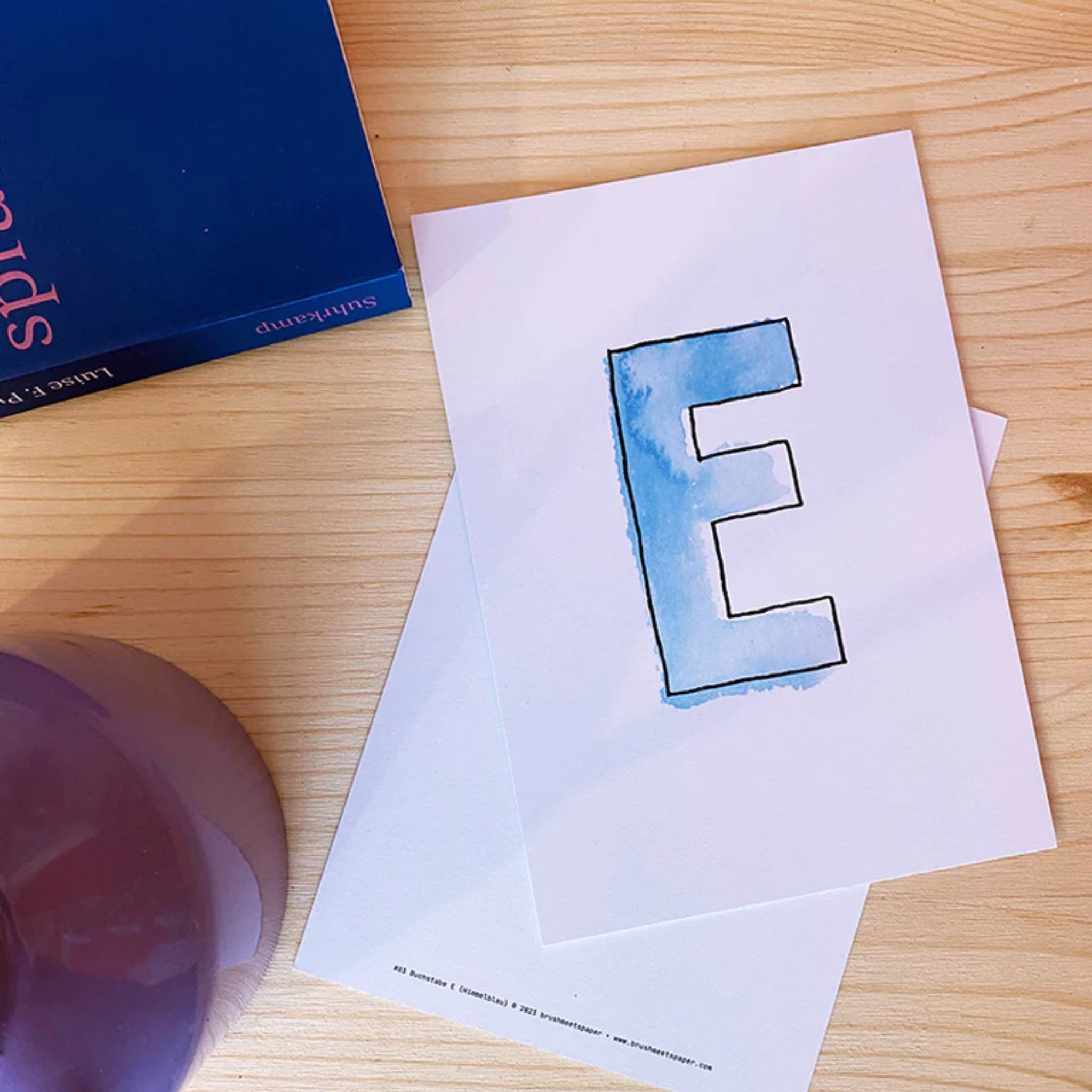 Ein Brief mit einem blauen, handgemalten Buchstaben "E" liegt auf einem Holztisch neben einem blauen Buch.