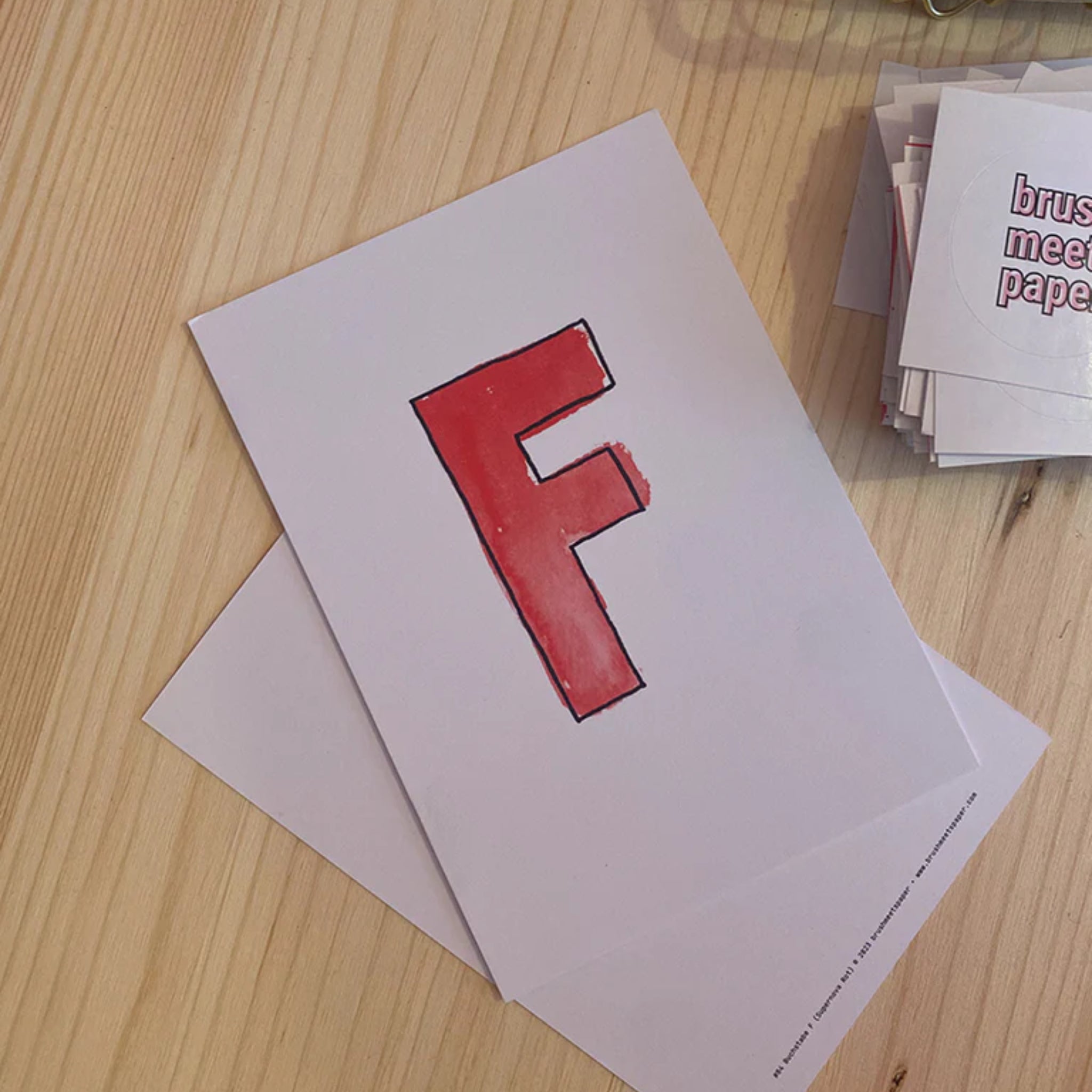 Ein Blatt Papier mit einem großen, roten Buchstaben „F“ liegt auf einem Holztisch neben einem Stapel anderer Papiere.