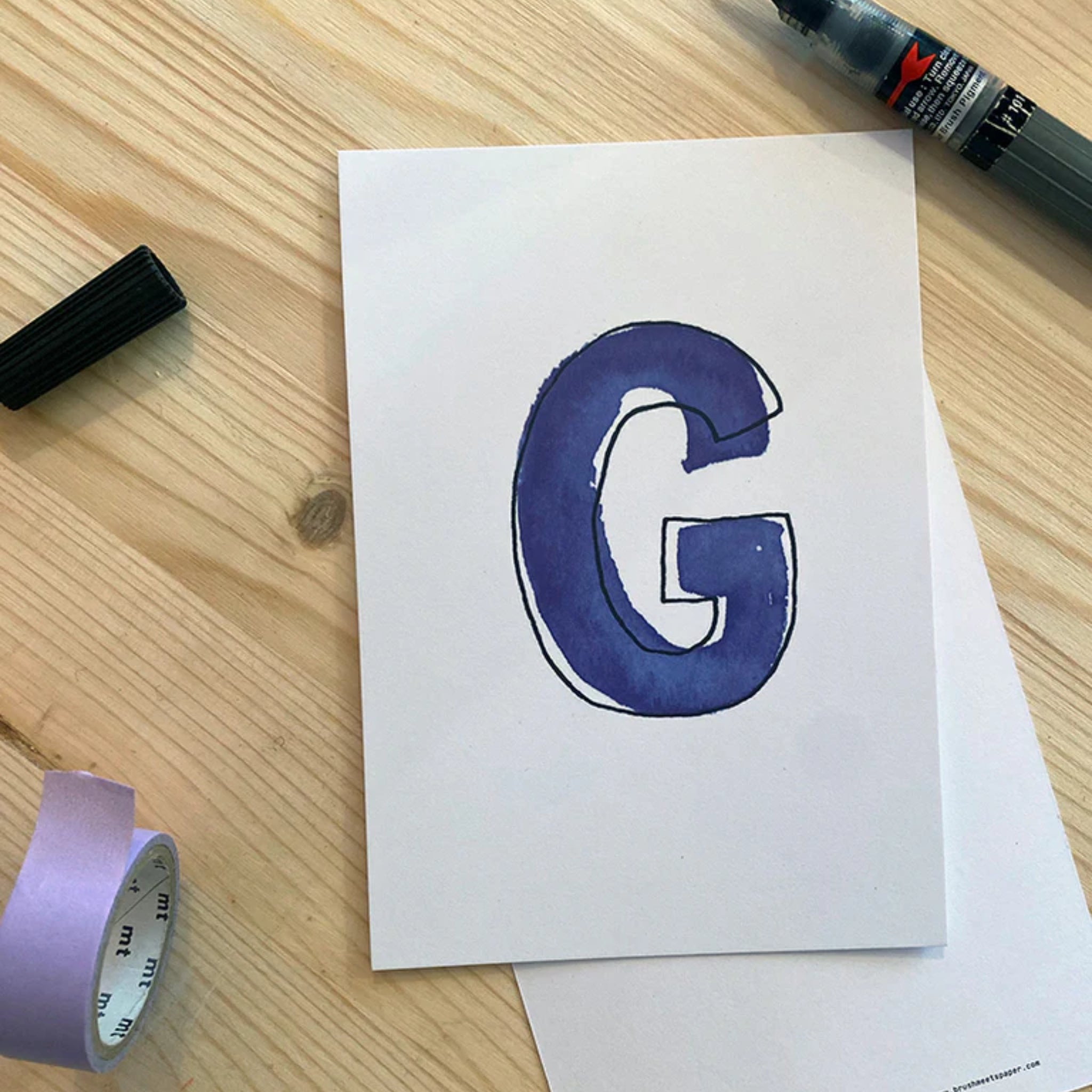 Pinselstift und lila Washi-Tape neben weißem Karton mit einem blauen Buchstaben "G" auf Holztisch.