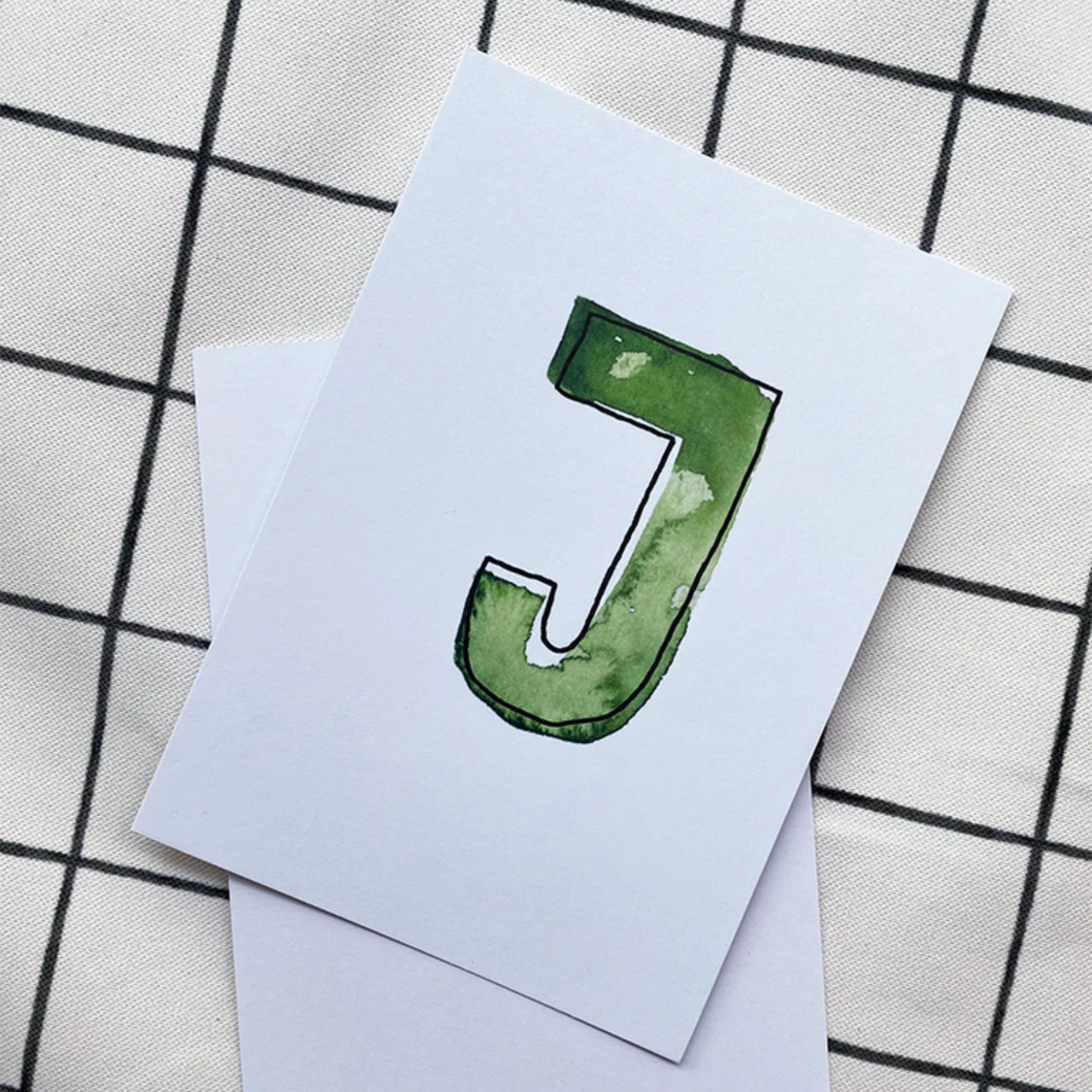 Weiße Karte mit einem großen, grünen Buchstaben "J" darauf, auf kariertem Hintergrund liegend.
