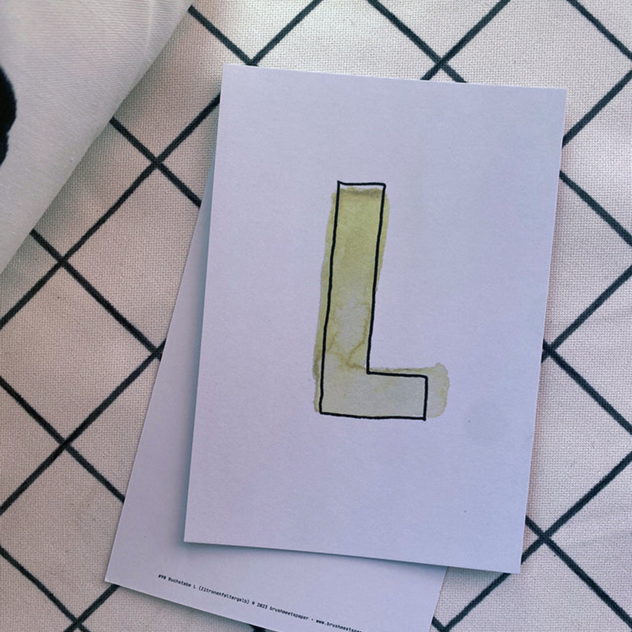 Eine weiße Karte mit einem gezeichneten, olivgrünen Buchstaben "L" liegt auf einem gitternetzförmigen Hintergrund.