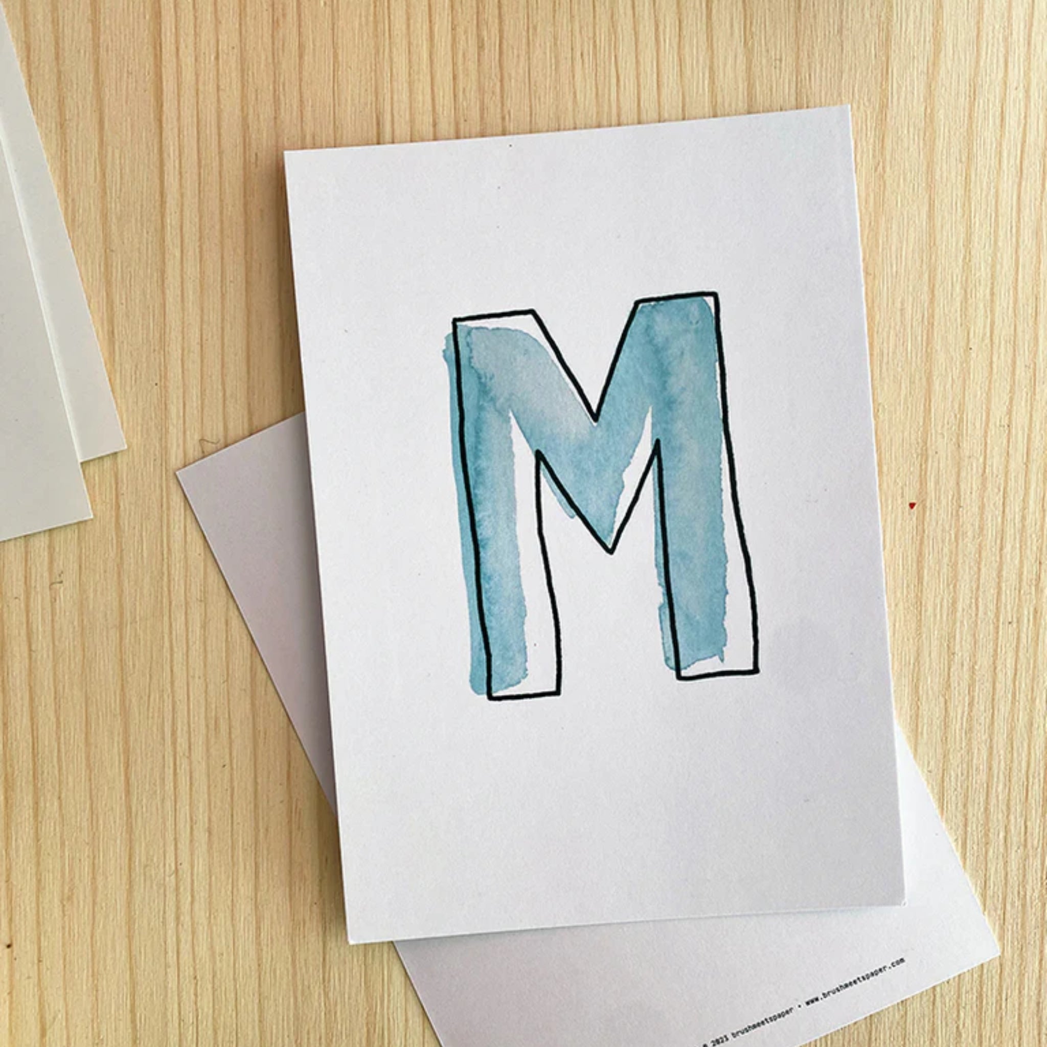 Weiße Karte mit großem, handgemaltem blauen Buchstaben "M" in Aquarelltechnik auf einem Holztisch.