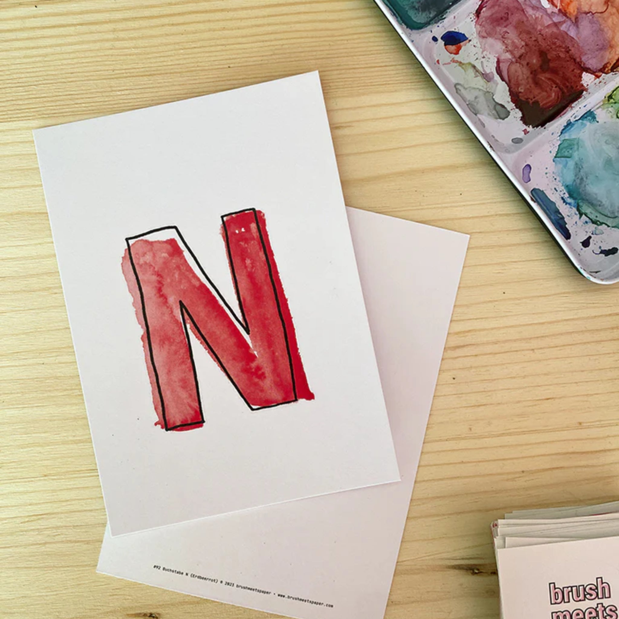 Eine handgemalte Karte mit einem roten Buchstaben "N" liegt auf einem Holztisch neben einer Farbpalette.