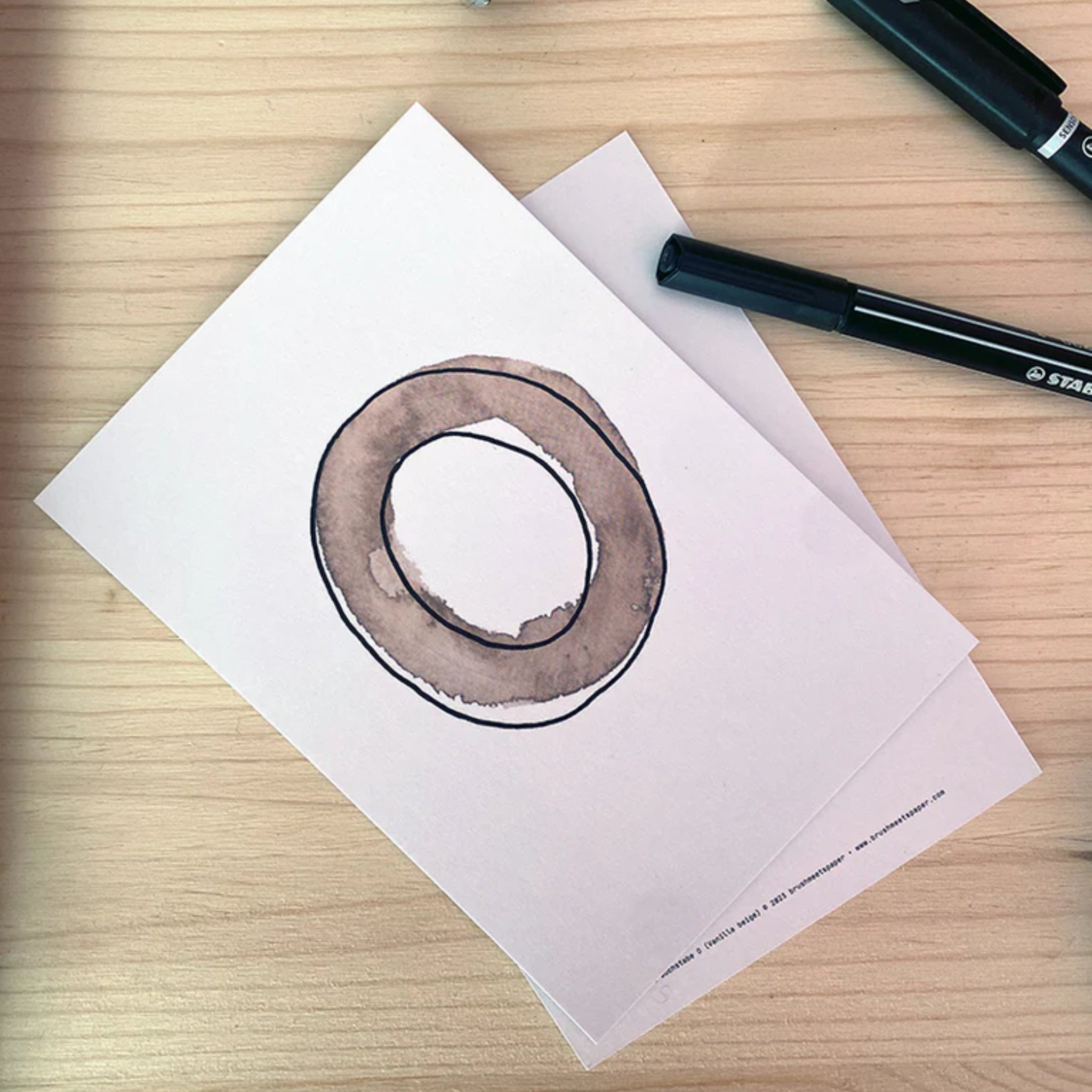 Ein handgezeichnetes braunes Donut-ähnliches O auf einem weißen Blatt Papier auf einem Holztisch neben zwei schwarzen Stiften.