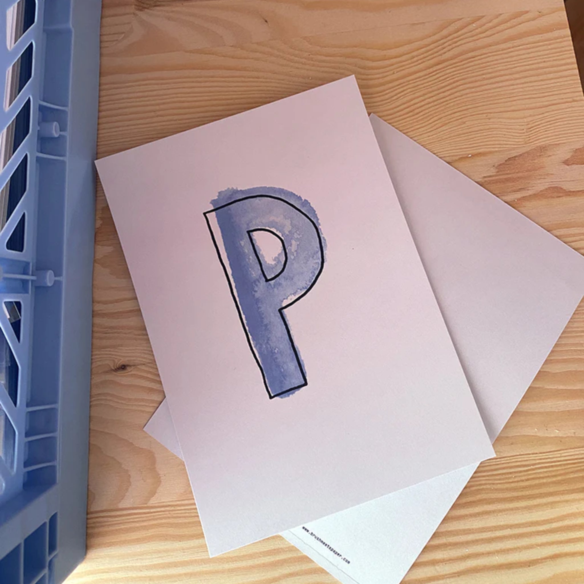Ein Blatt Papier mit einem blauen, handgemalten, großen Buchstaben "P" liegt auf einem Tisch neben einem blauen Fach.