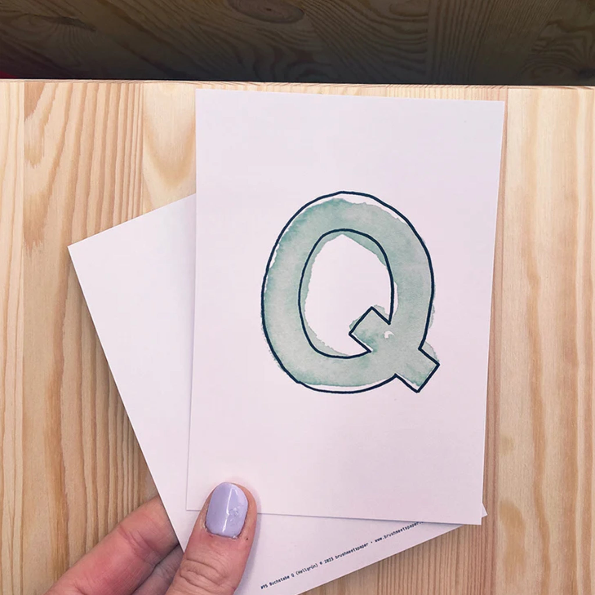 Eine Hand hält eine Karte mit einem großen, grün-aquarellierten Buchstaben "Q" auf einem Holztisch.