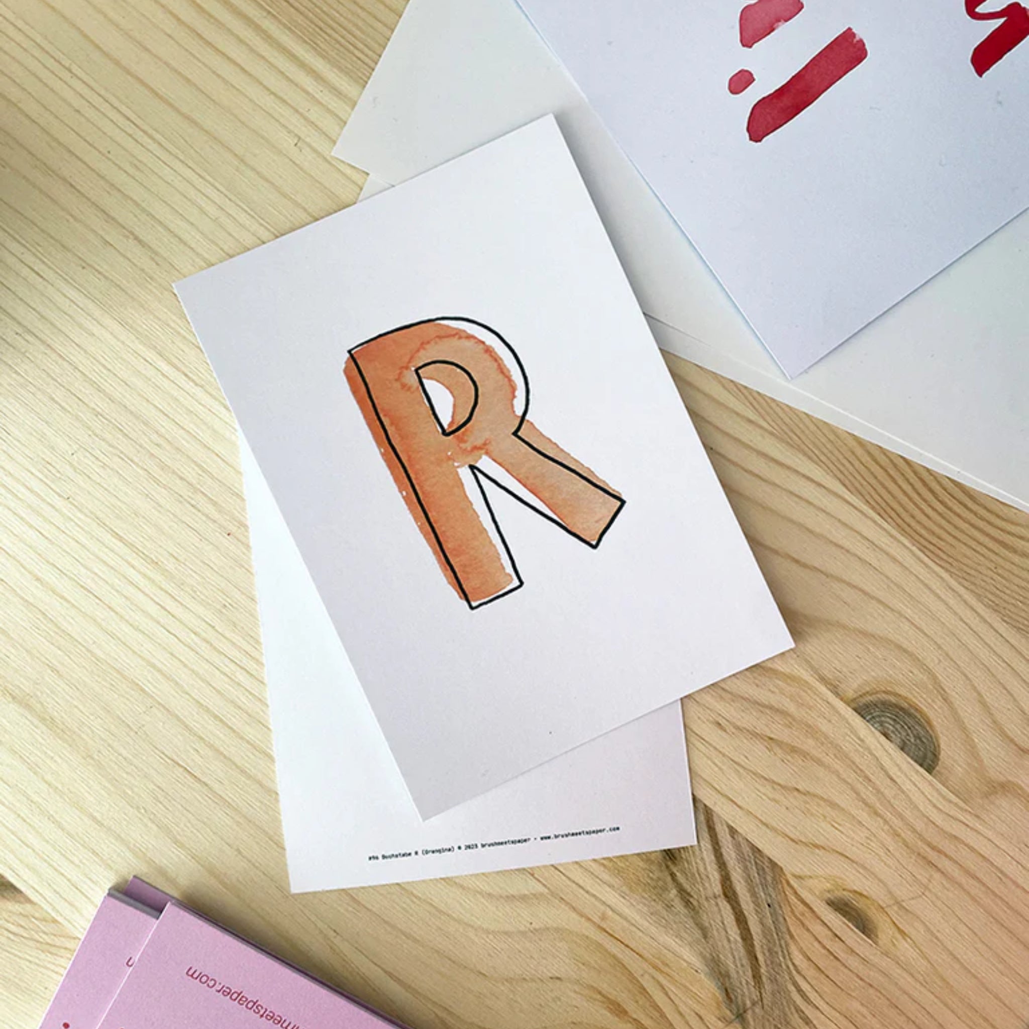 Ein Blatt Papier mit einem handgemalten, orangen Buchstaben "R" liegt auf einem Holztisch.