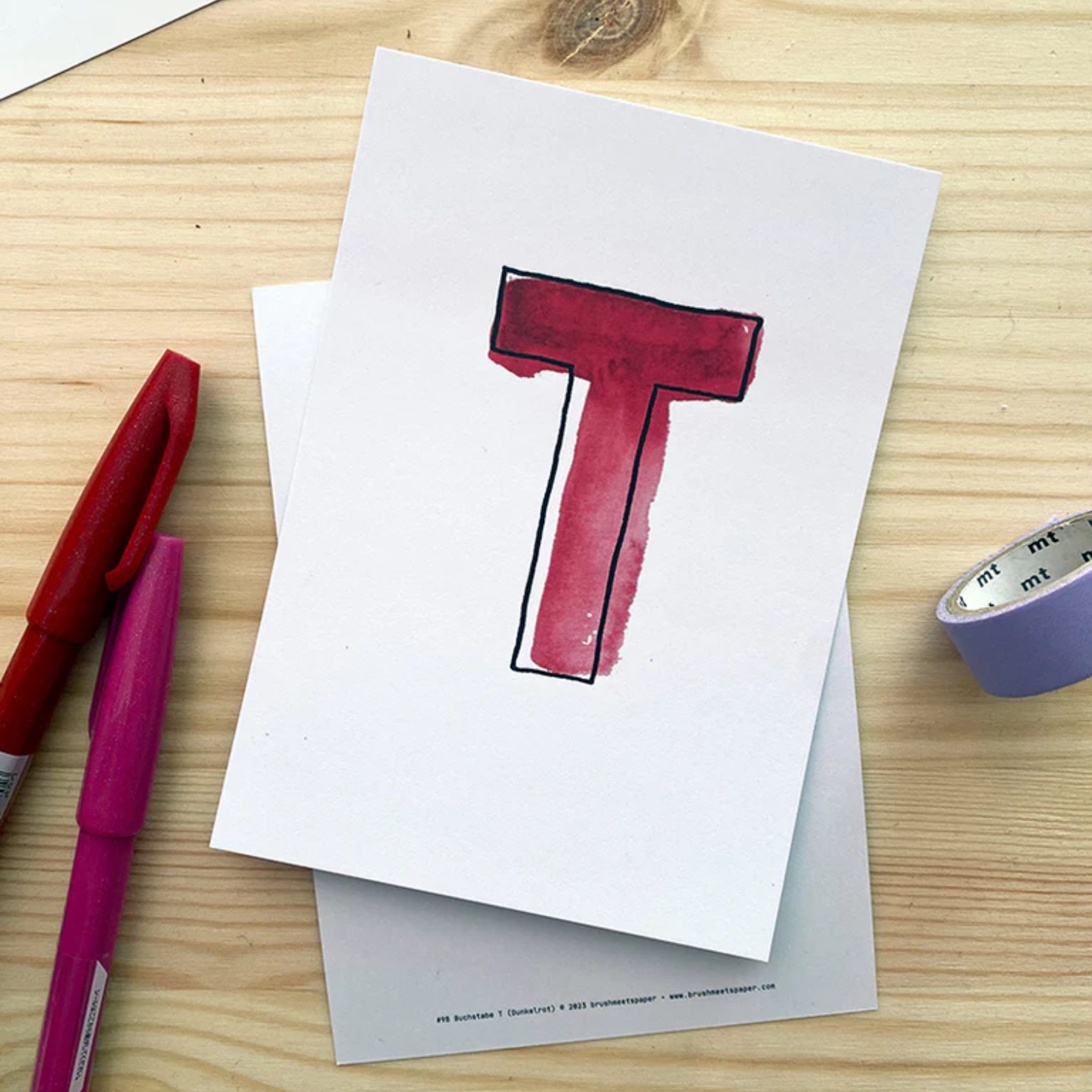 Ein handgemaltes rotes "T" auf einer weißen Karte liegt auf einem Holztisch, umgeben von Stiften und Klebeband.
