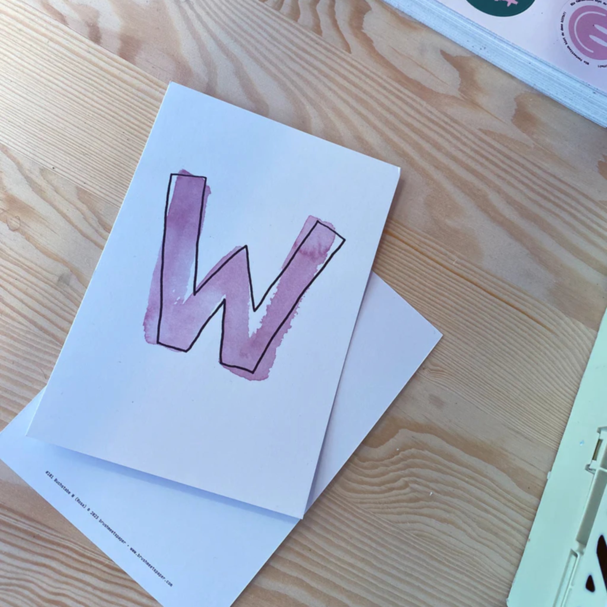 Ein weißes Blatt Papier mit einem handgemalten, lila Buchstaben "W" liegt auf einem Holztisch.