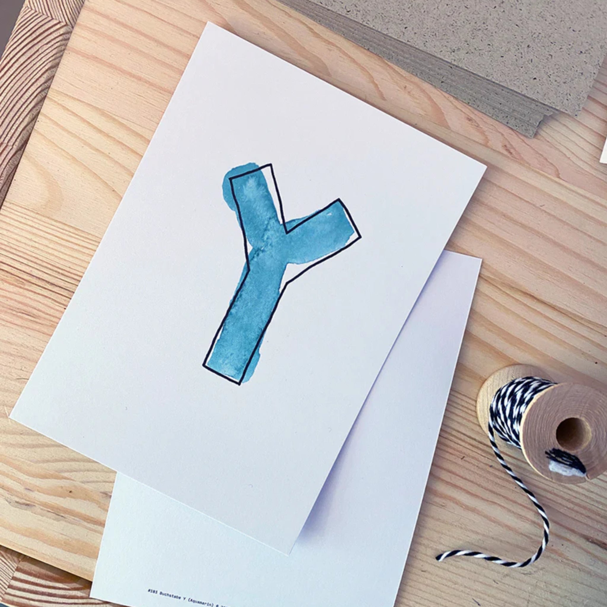 Ein blauer Buchstabe "Y" auf weißem Papier liegt auf einem Holztisch neben einer Rolle mit schwarz-weißem Garn.