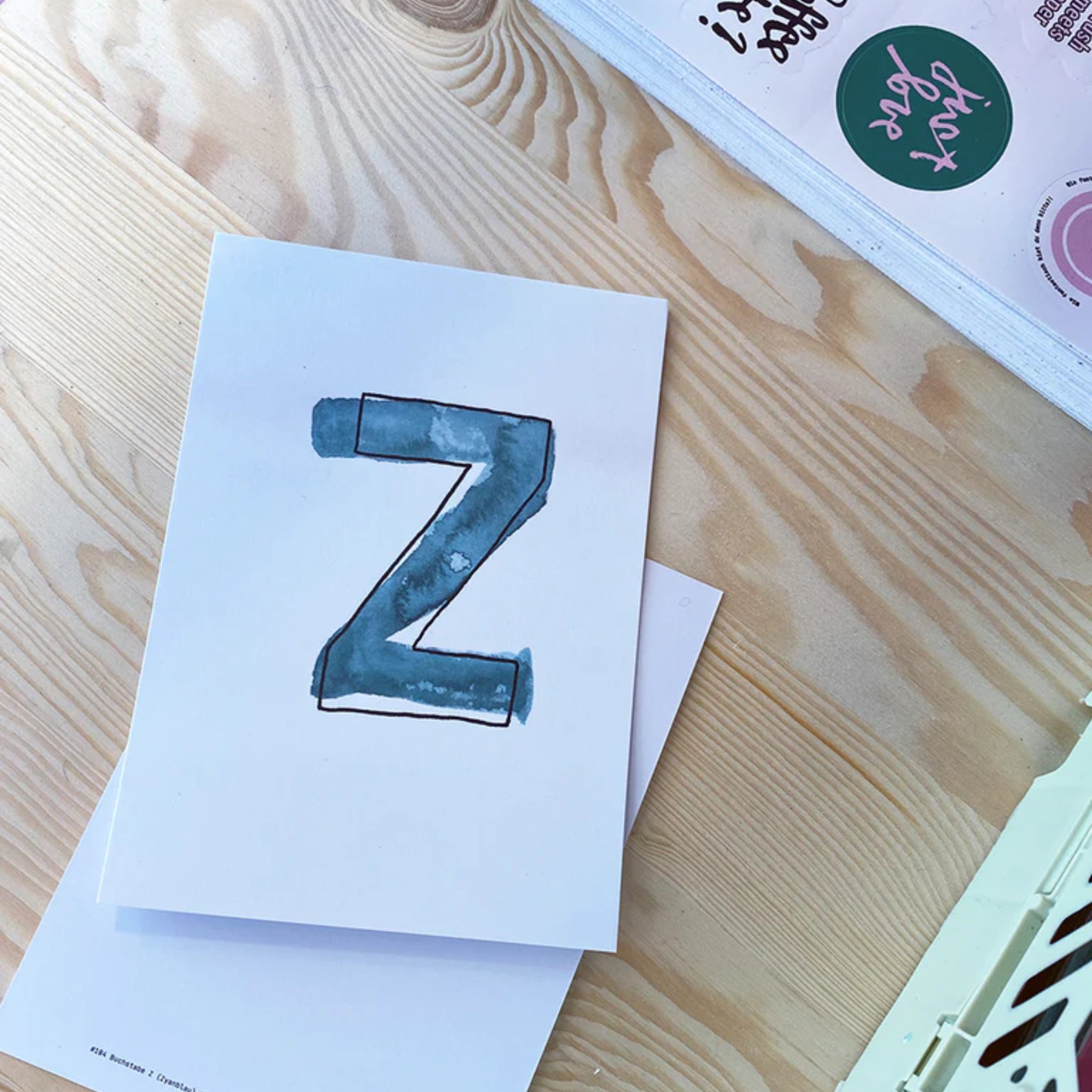 Ein weißes Blatt mit einem blauen, gemalten Buchstaben "Z" liegt auf einem hellen Holztisch.
