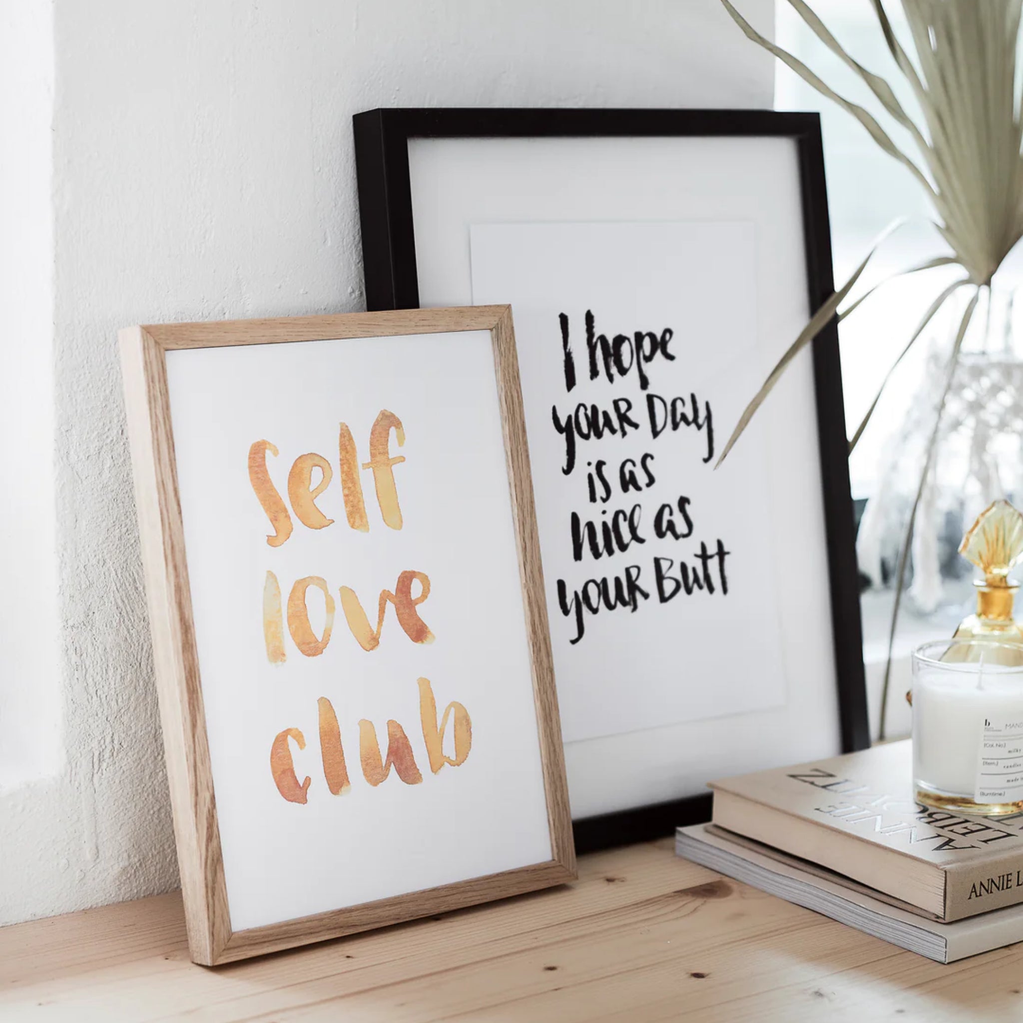 Zwei gerahmte Zitate auf einem Tisch. Links: "Self Love Club", rechts: "I hope your day is as nice as your butt".