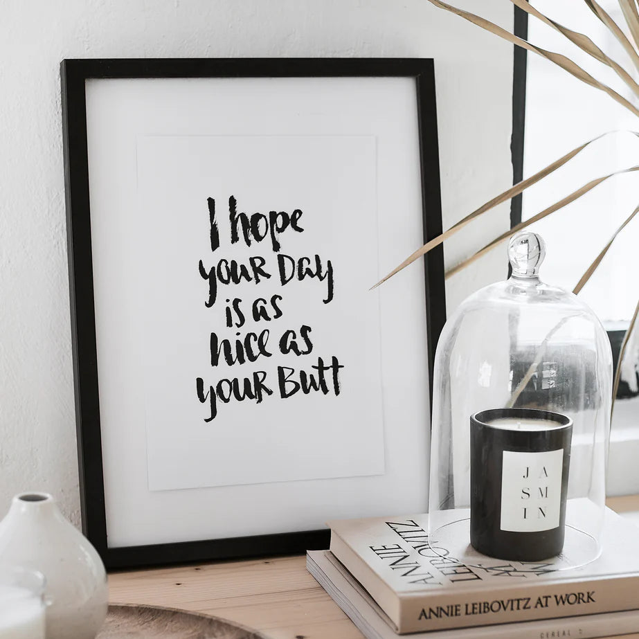 Gerahmter Druck mit der Aufschrift "I hope your day is as nice as your butt" neben einer Kerze auf gestapelten Büchern.