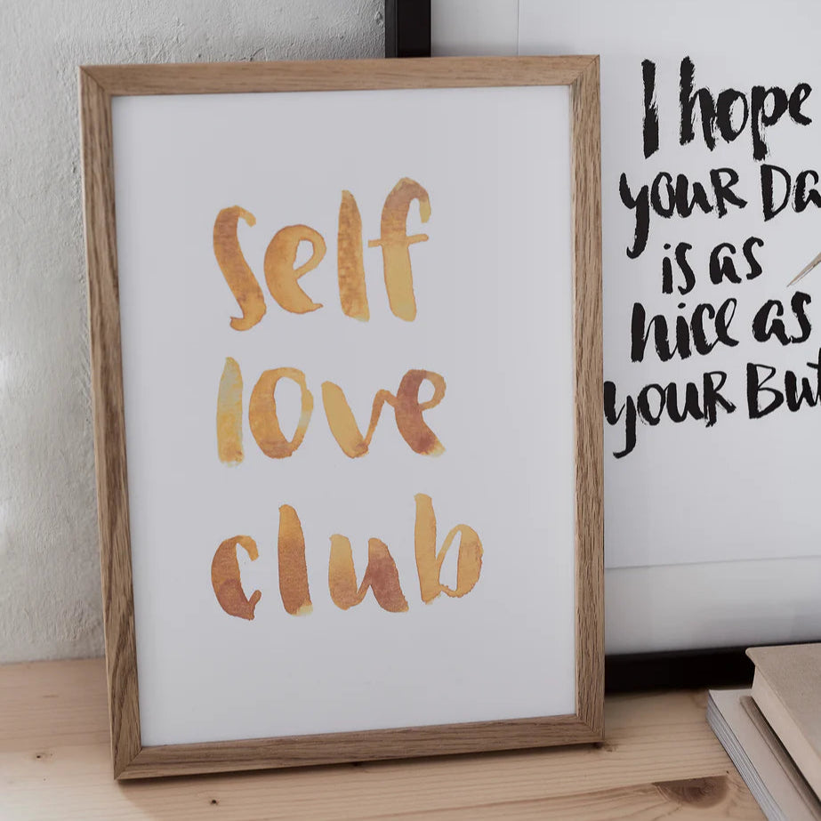 Holzgerahmtes Poster mit der Aufschrift "self love club" in orange Aquarellschrift auf einem Tisch angelehnt.