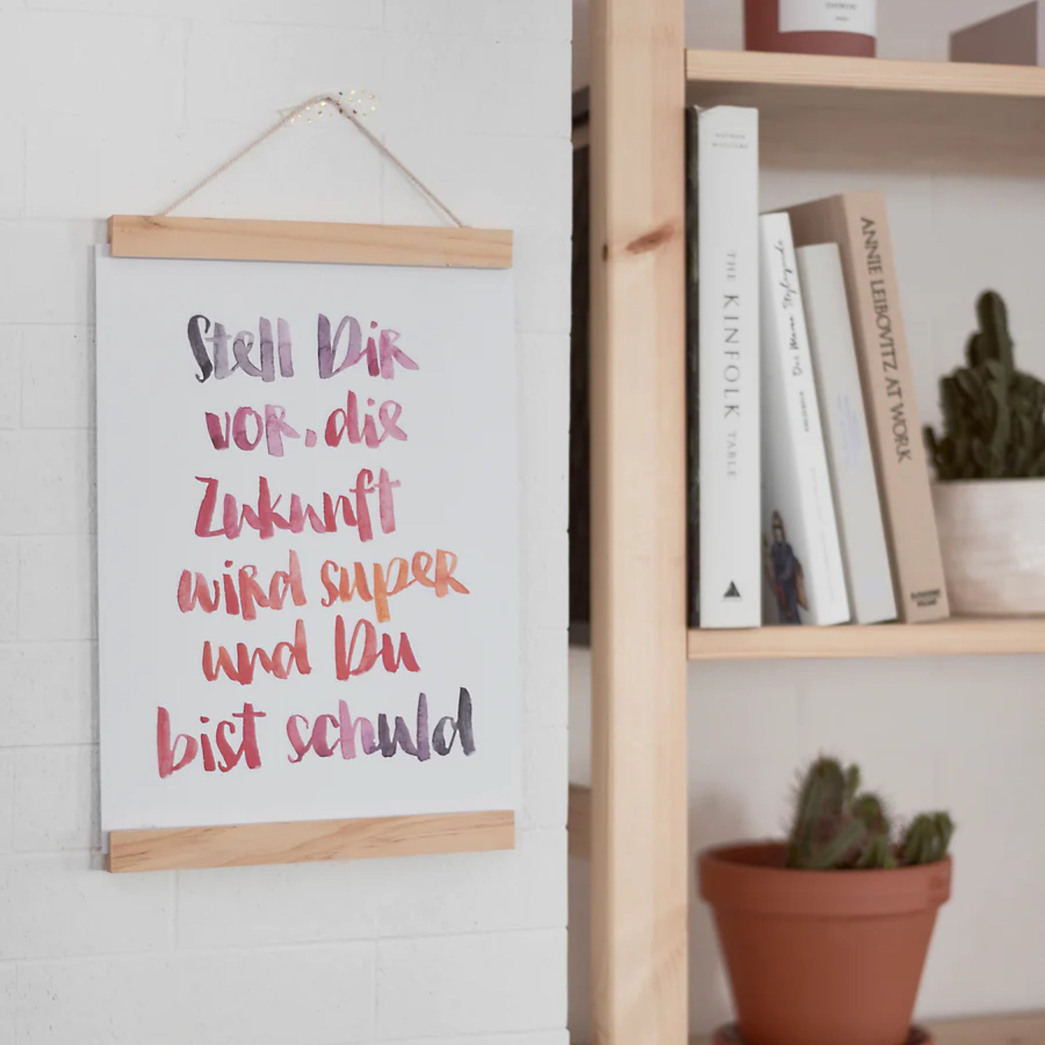 Poster mit Schriftzug auf Deutsch hängt neben einem Bücherregal mit Kaktus und Büchern.