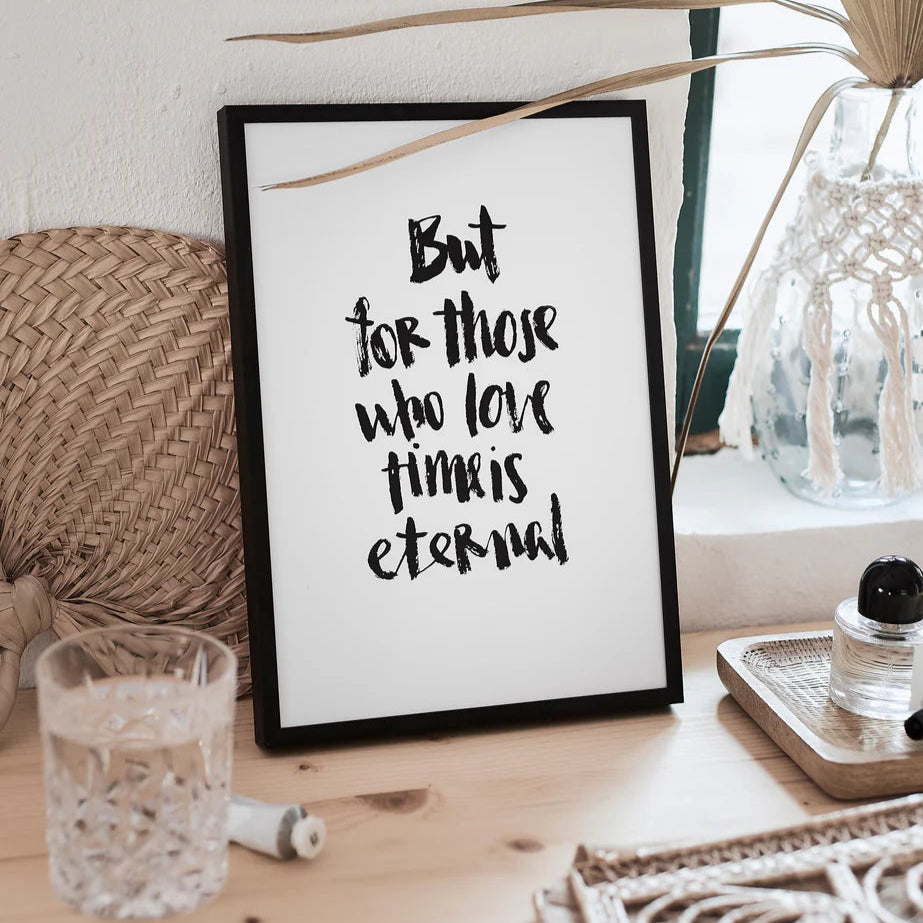 Gerahmtes Zitatbild auf Holztisch mit Glas Wasser und Dekoration, das den Text "But for those who love time is eternal" zeigt.