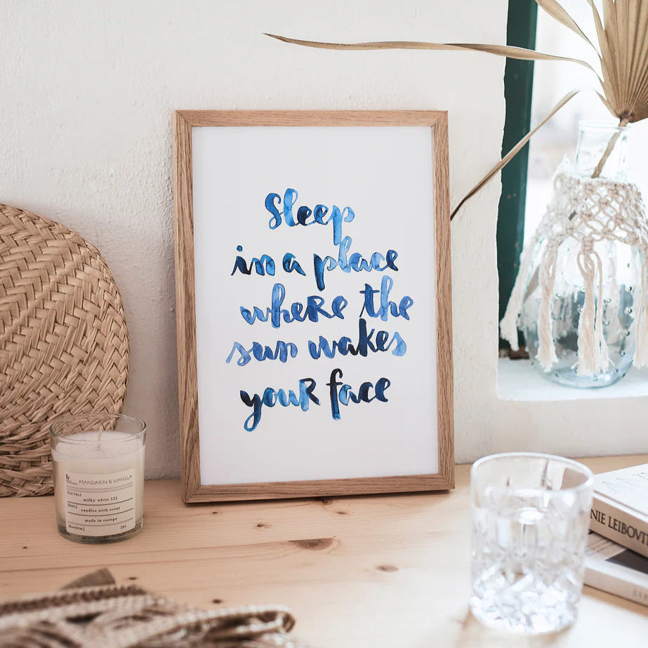 Ein gerahmtes Bild mit dem Text "Sleep in a place where the sun wakes your face" steht auf einem Tisch neben Kerze und Glas.