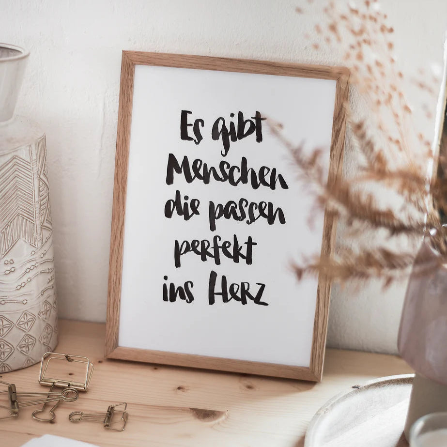 Gerahmter Spruch: "Es gibt Menschen die passen perfekt ins Herz" auf einem Tisch mit Dekorationen.