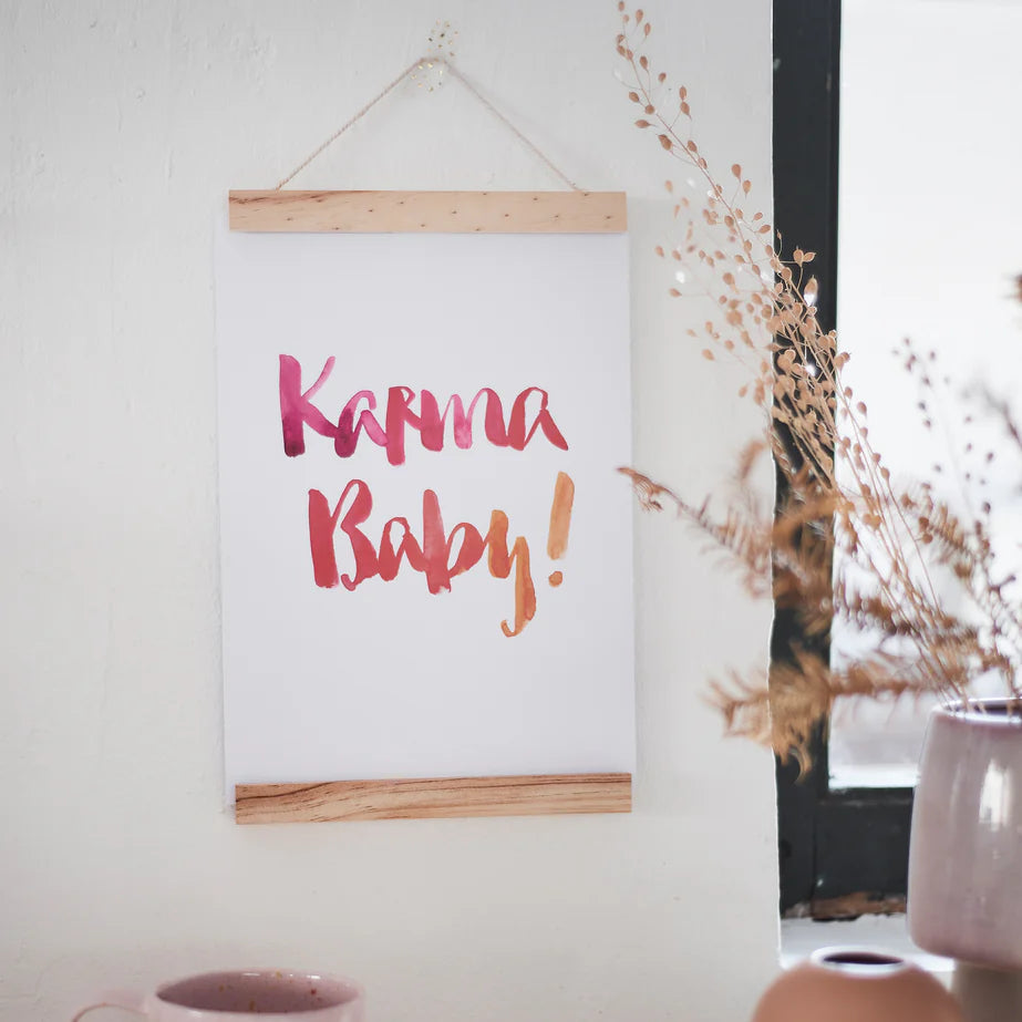 Ein Poster mit der Aufschrift "Karma Baby!" hängt an einer Wand neben einer Fensterbank mit Trockenblumen.