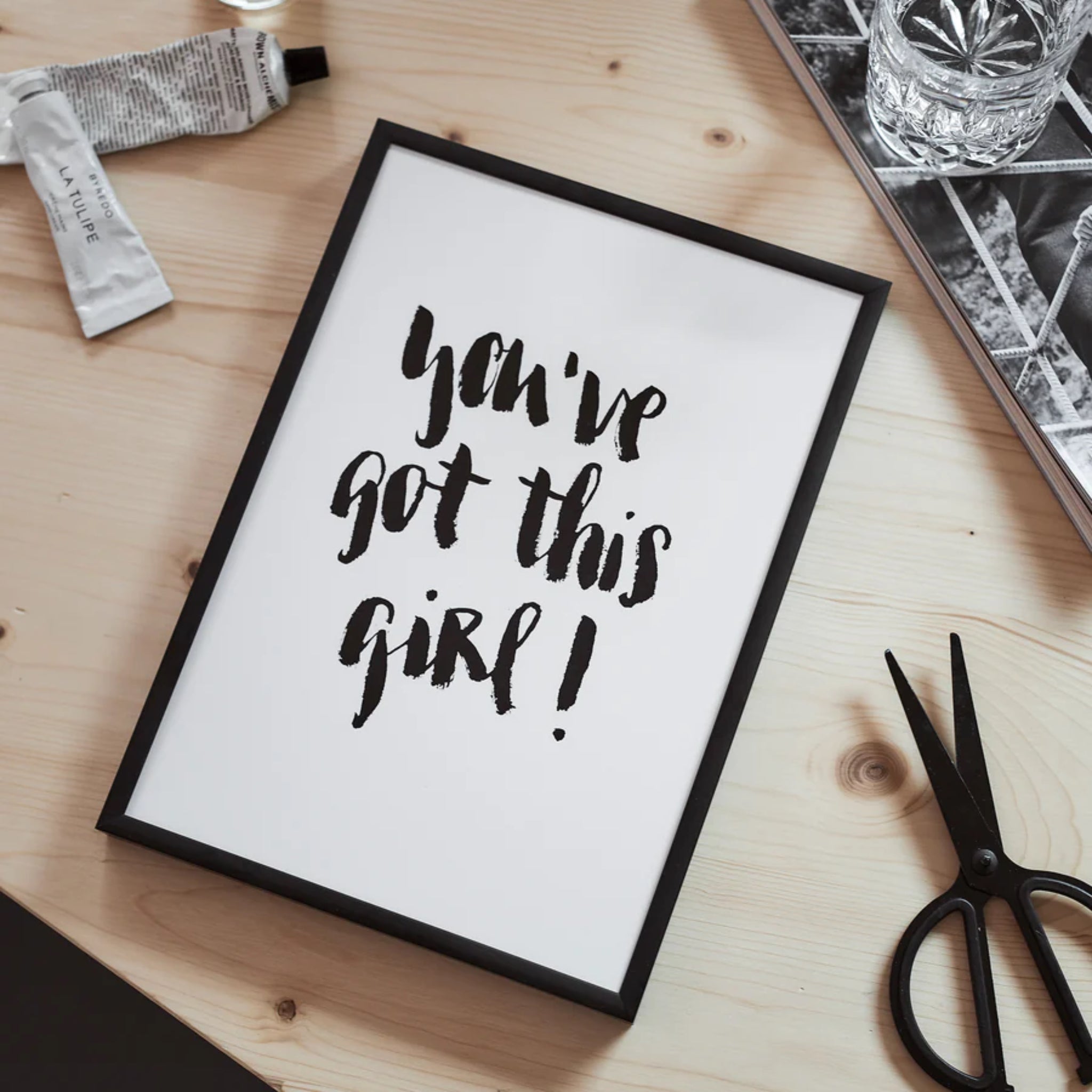 Rahmen mit der Aufschrift "You've got this girl!" auf einem Tisch neben Schere und einem Glas.